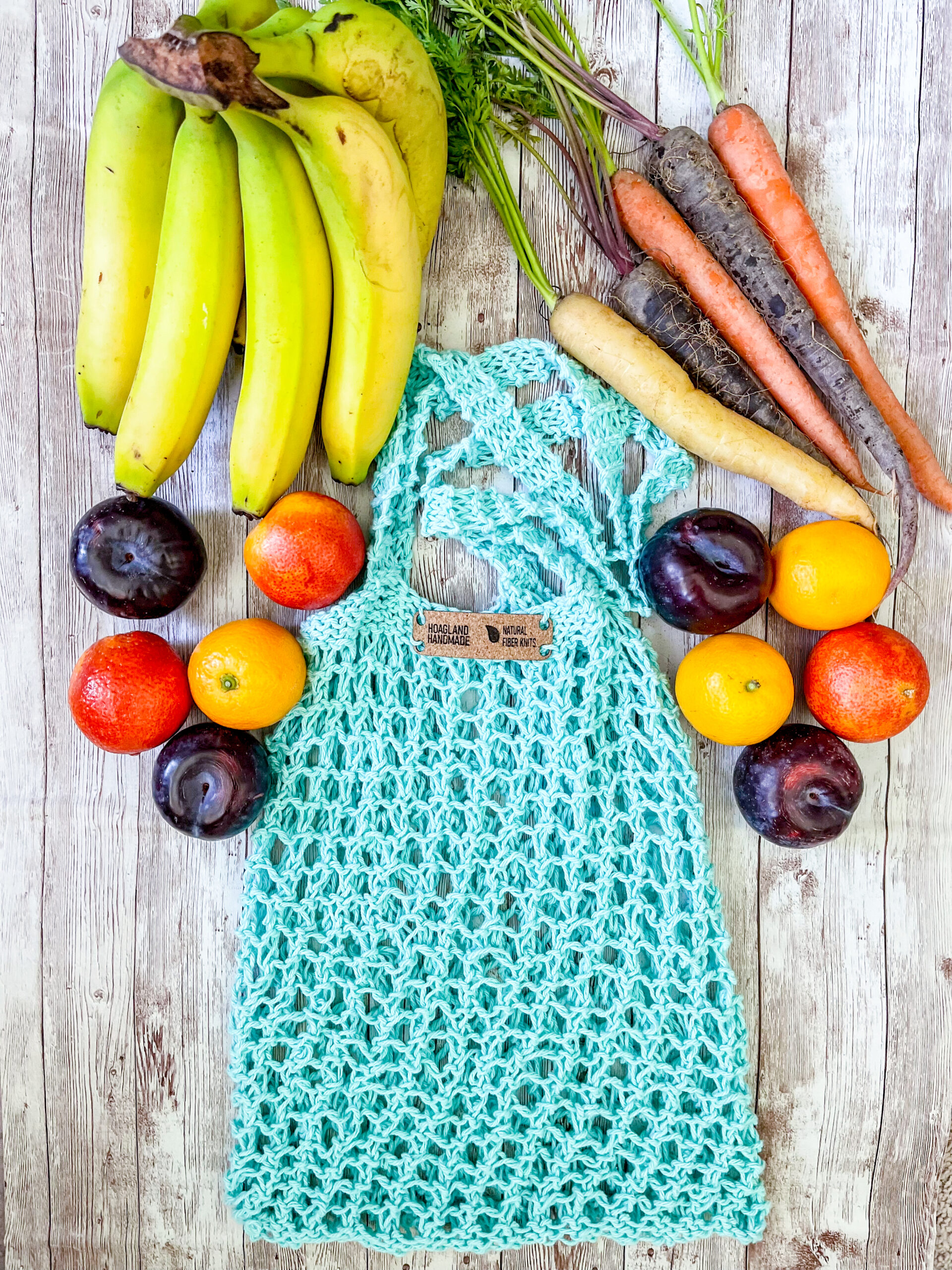 A teal recycled cotton mesh tote bag is surrounded by bananas, rainbow carrots, plums, oranges, and blood oranges