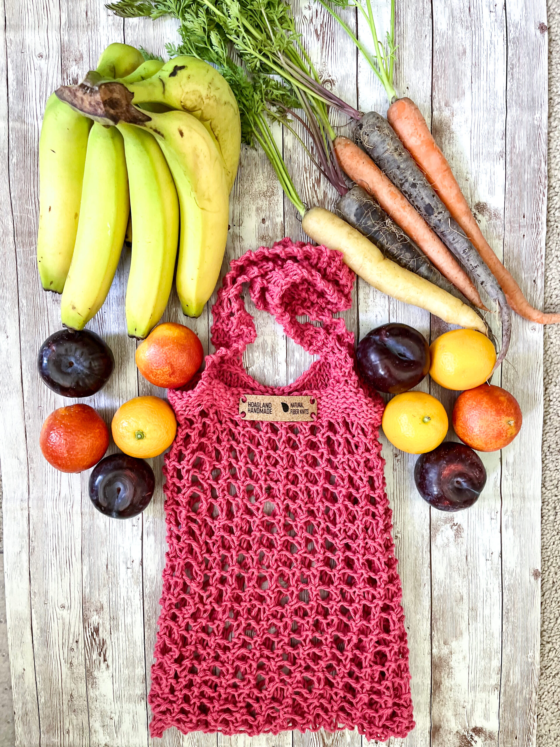 A pink recycled cotton mesh tote bag is surrounded by bananas, rainbow carrots, plums, oranges, and blood oranges