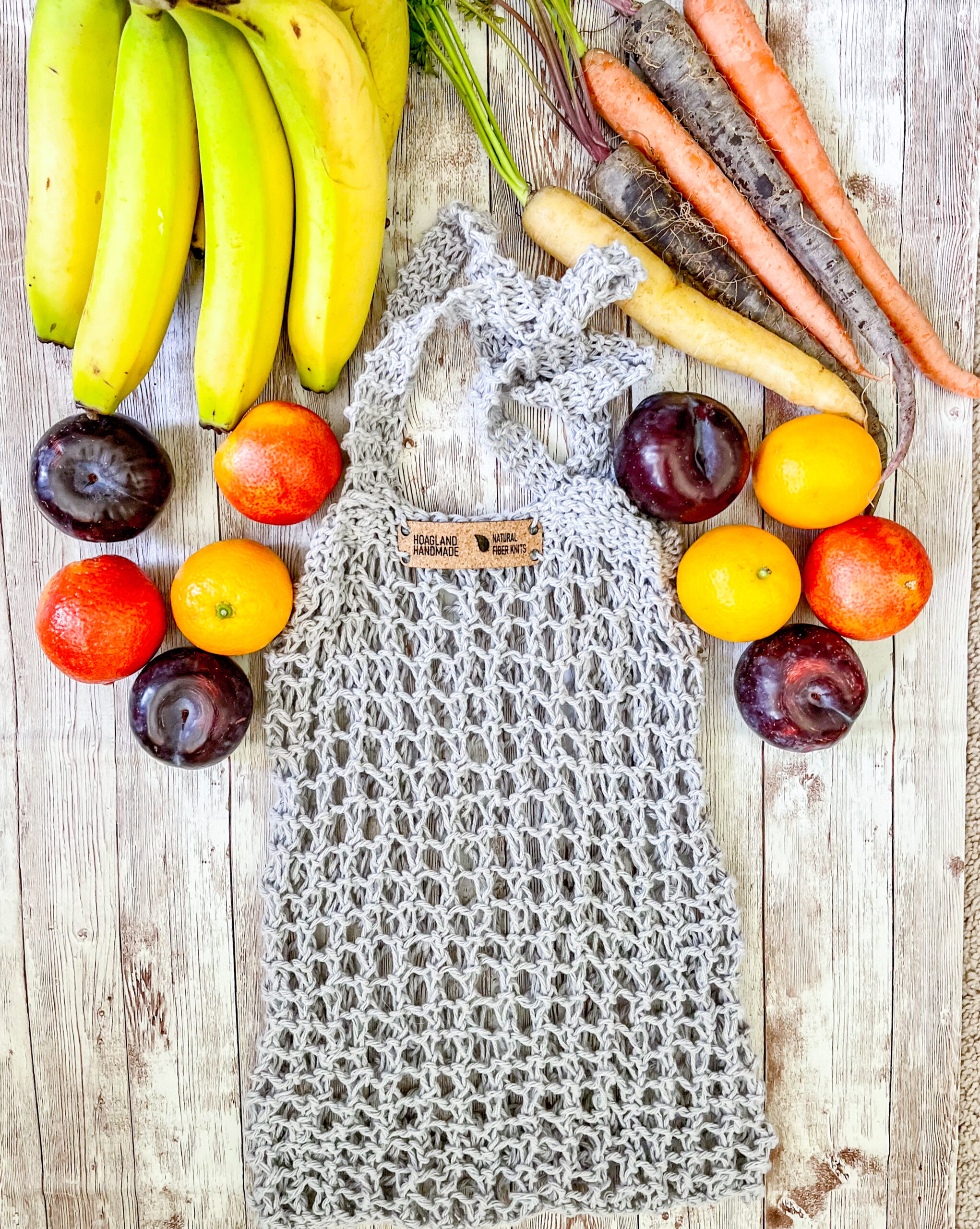 A gray recycled cotton mesh tote bag is surrounded by bananas, rainbow carrots, plums, oranges, and blood oranges