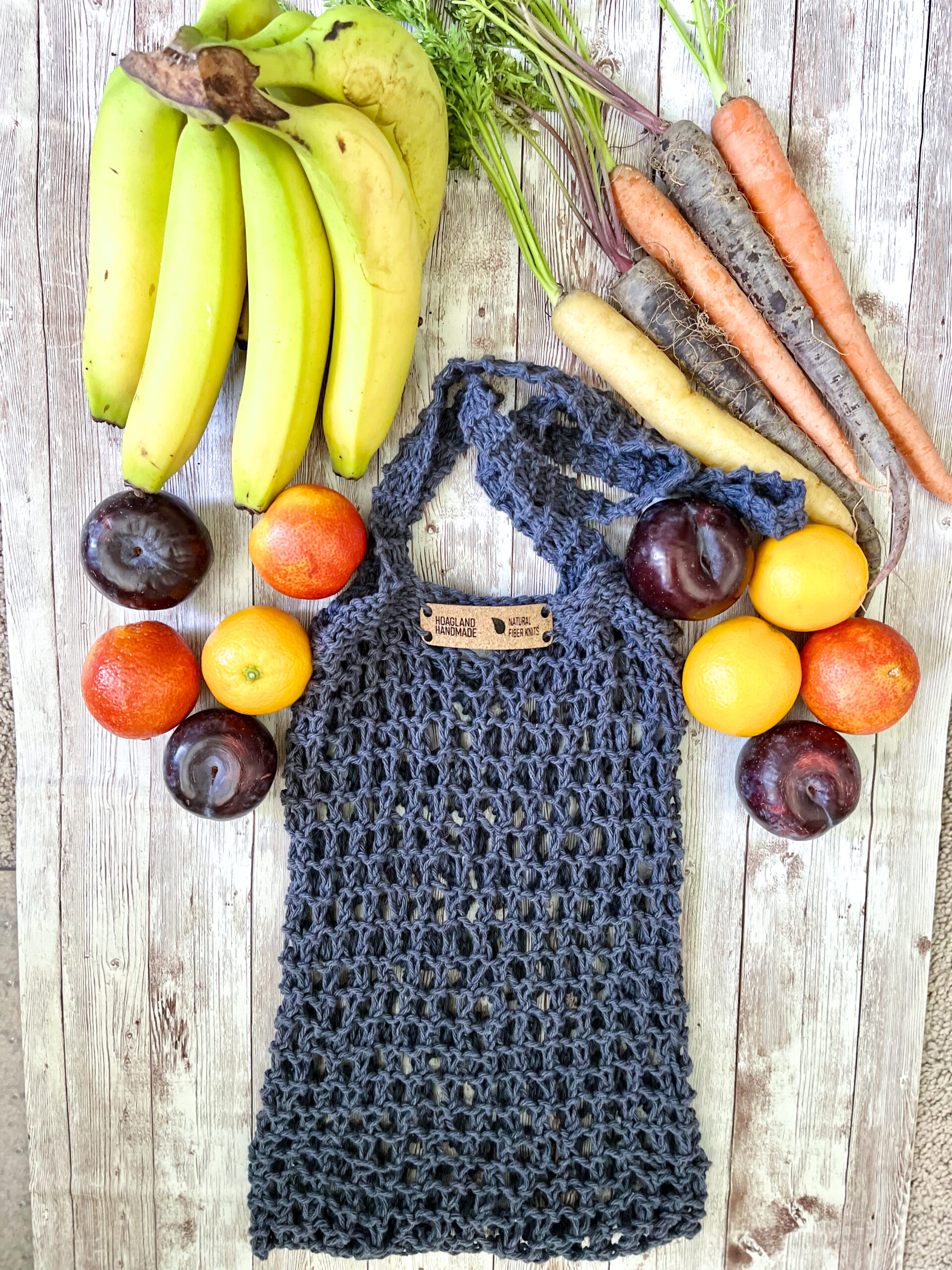 A dark blue recycled cotton mesh tote bag is surrounded by bananas, rainbow carrots, plums, oranges, and blood oranges