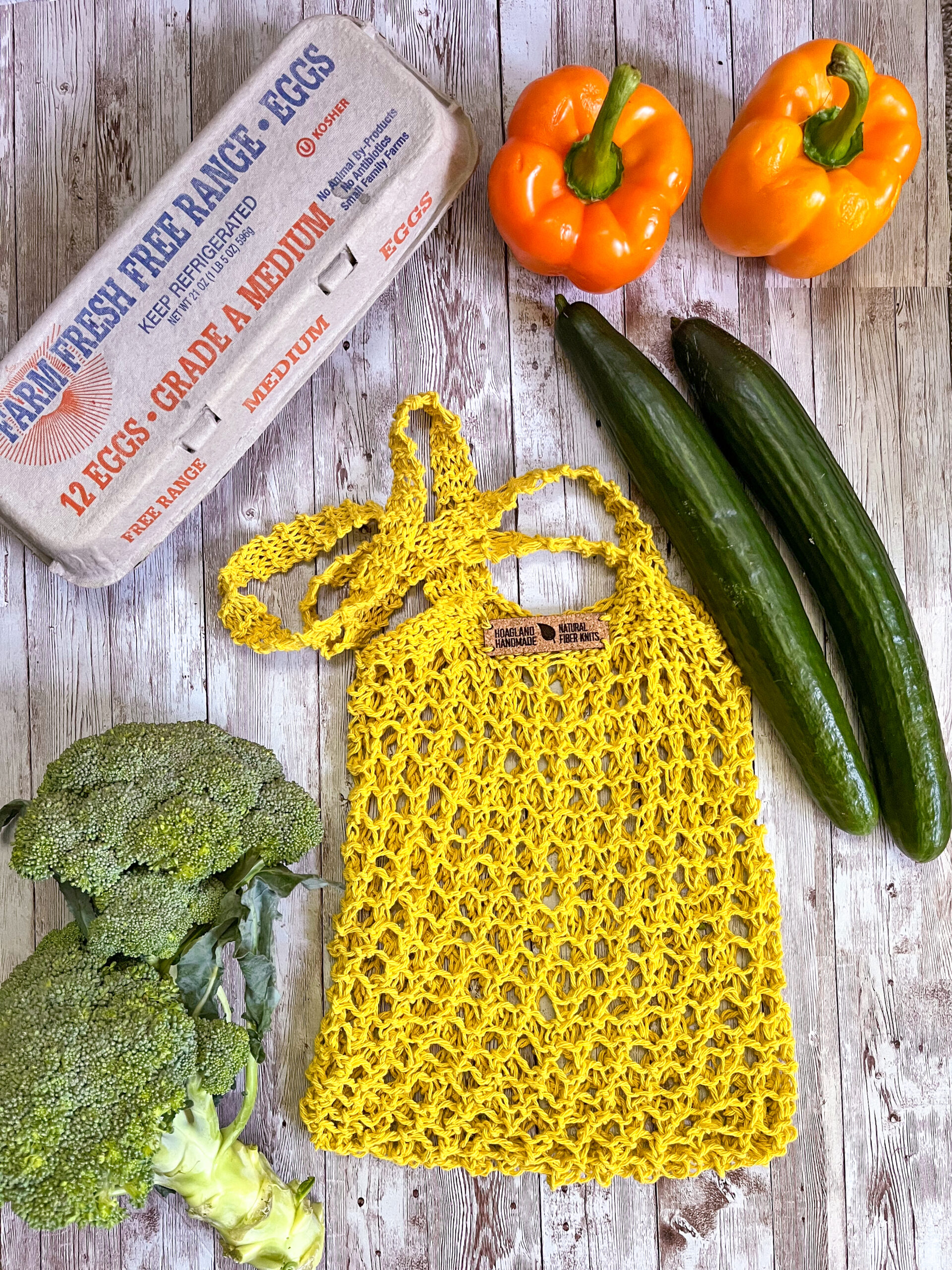 A yellow recycled cotton mesh tote bag is surrounded by a carton of farm eggs, orange bell peppers, cucumbers, and broccoli