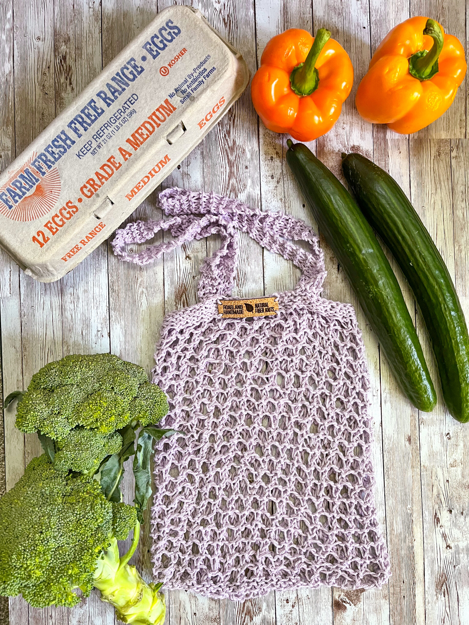 A purple recycled cotton mesh tote bag is surrounded by a carton of farm eggs, orange bell peppers, cucumbers, and broccoli