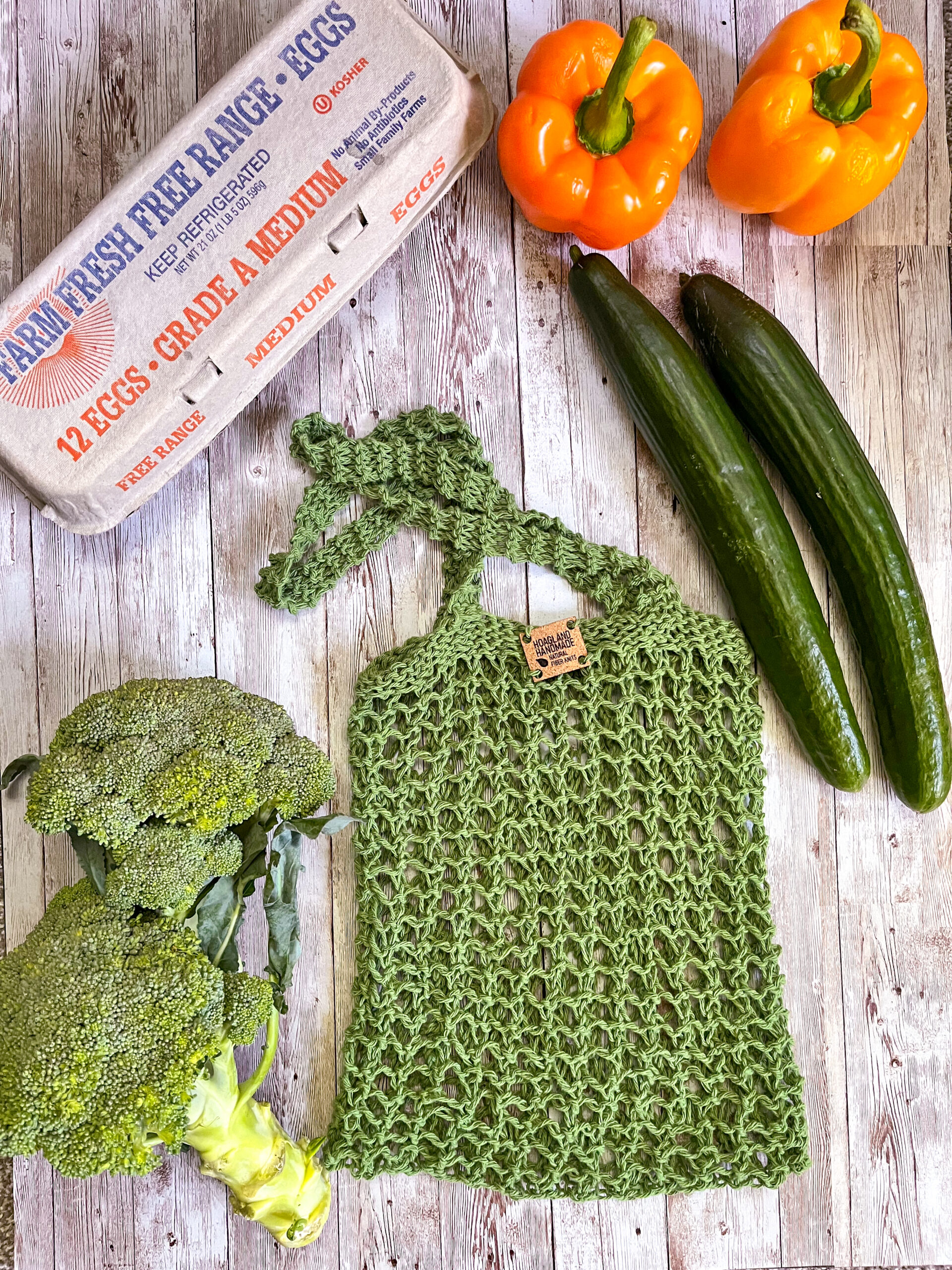 A green recycled cotton mesh tote bag is surrounded by a carton of farm eggs, orange bell peppers, cucumbers, and broccoli