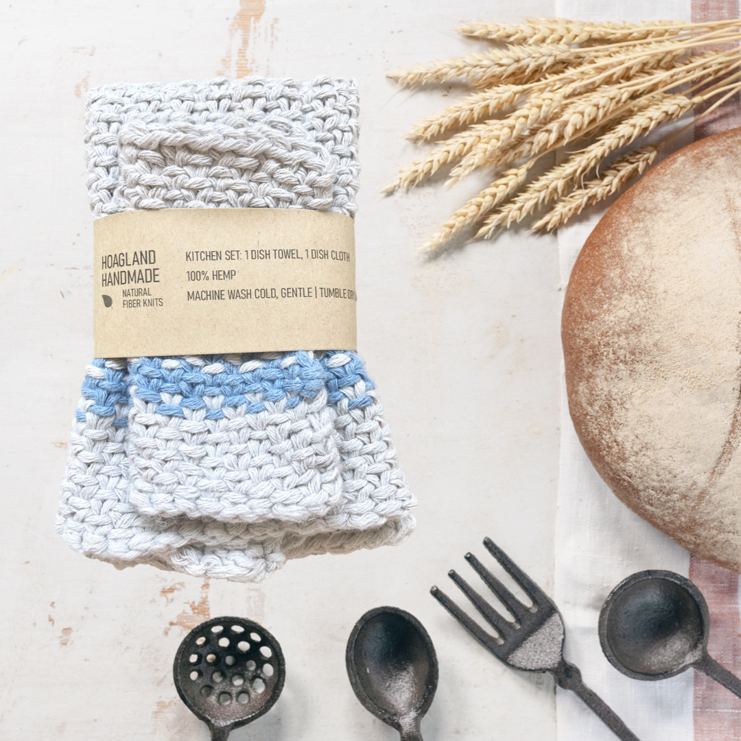 A wrapped natural with blue stripe dish towel set sits next to a loaf of homemade bread, a bunch of wheat, and several kitchen utensils