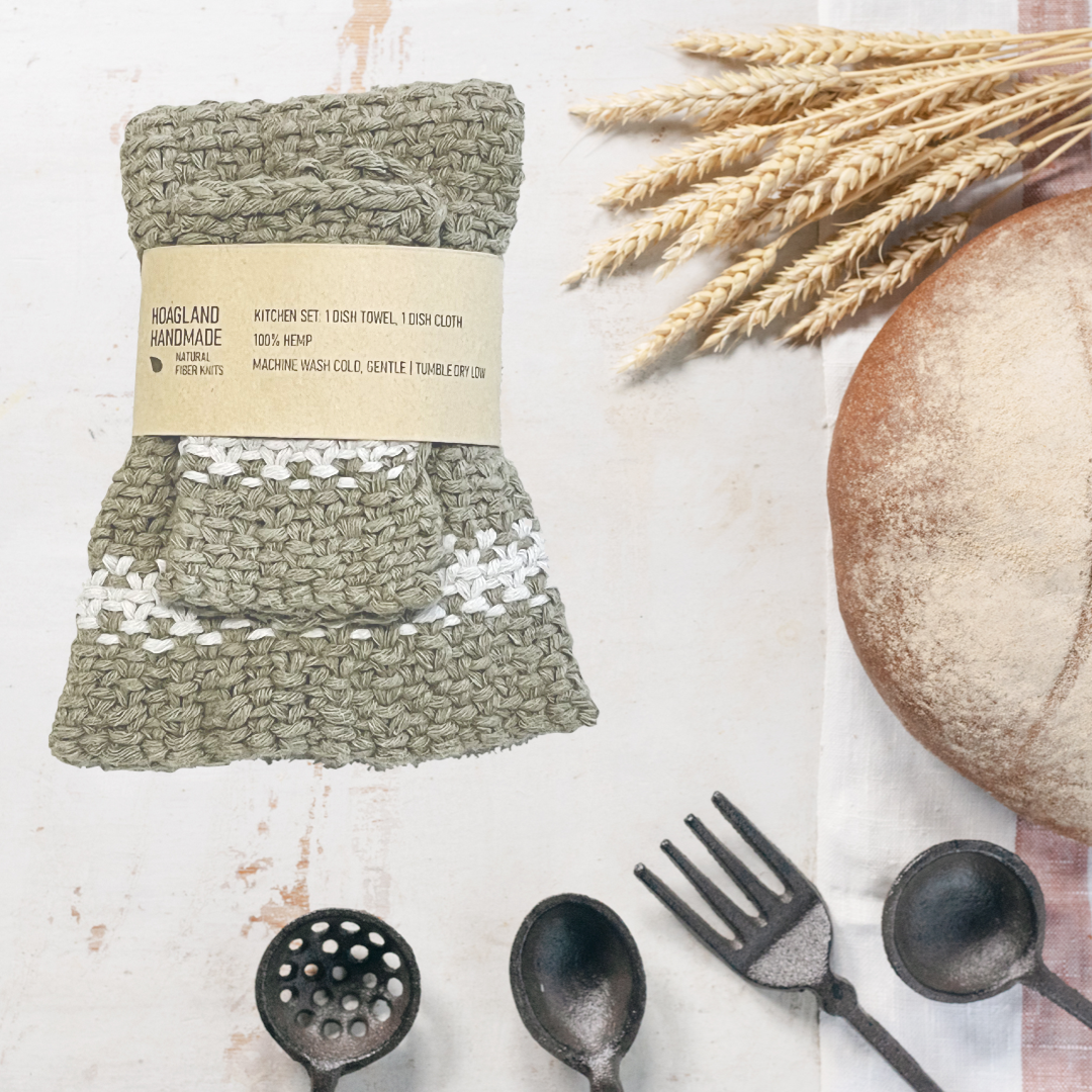 A wrapped green with natural white stripe dish towel set sits next to a loaf of homemade bread, a bunch of wheat, and several kitchen utensils