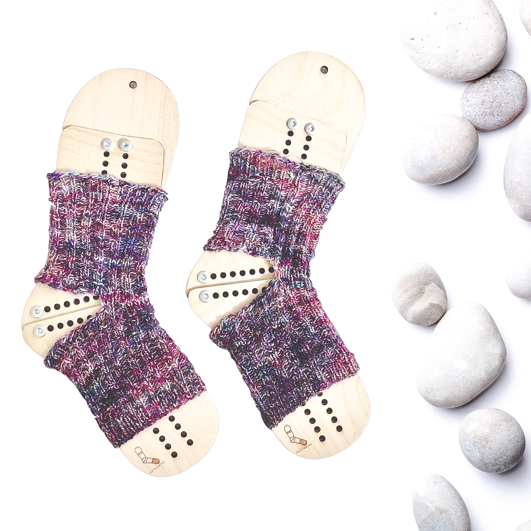 A pair of speckled yoga socks are displayed on a set of wooden sock blockers with gray rocks along the right side of the photo. The yoga socks are a mix of purples, blues, reds, and pinks