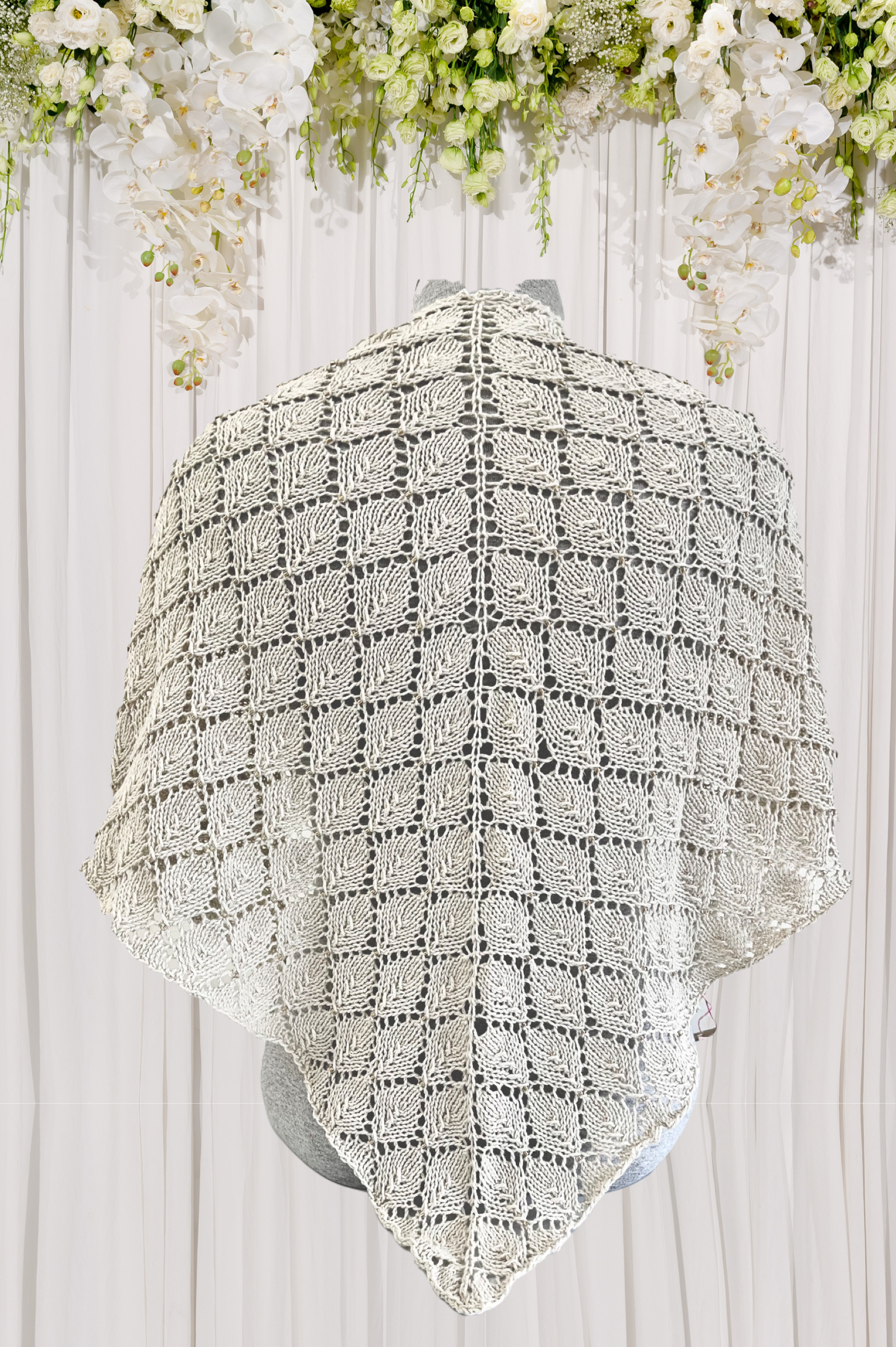 An organic merino/cotton beaded shawl is hand knit in a stunning leaf lace pattern, with subtle coordinating Czech glass beads. It is shown over a wedding dress, with a white fabric draping in the background with white flowers draped over it.