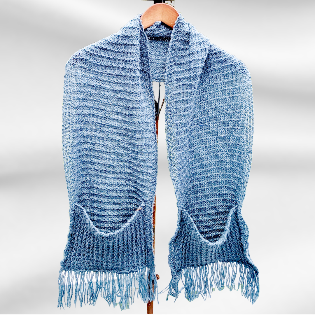 A medium blue scarf hangs from a hanger on a stand. It is an oversized scarf with pockets and fringe on each end
