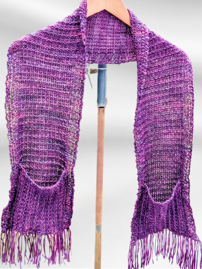 A hand dyed merino purple scarf hangs draped around a hanger. The scarf has oversized pockets and fringe on each end