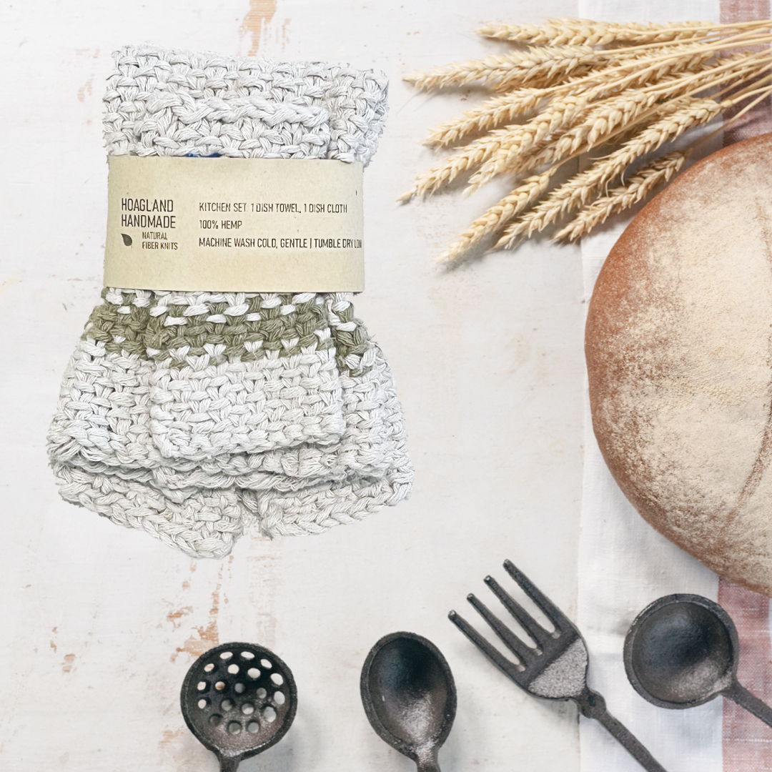 A wrapped natural with green stripe dish towel set sits next to a loaf of homemade bread, a bunch of wheat, and several kitchen utensils