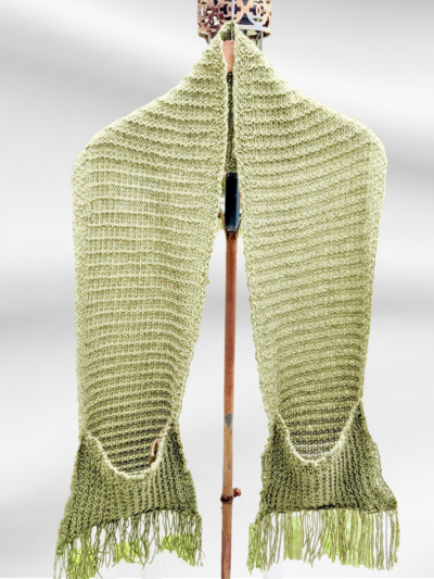 A mossy green scarf hangs from a hanger on a stand. It is an oversized scarf with pockets and fringe on each end