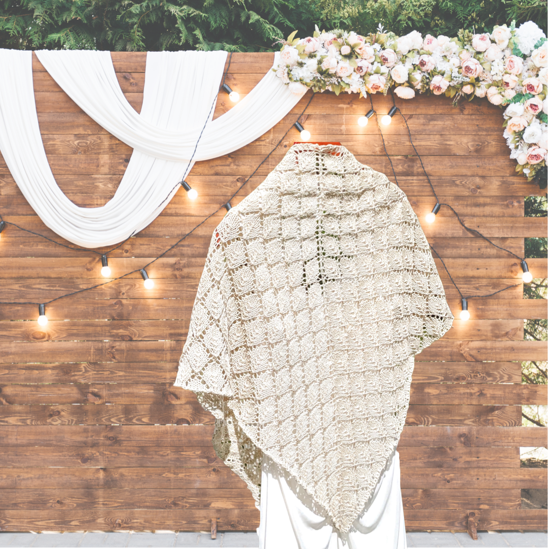 An organic merino/cotton beaded shawl is hand knit in a stunning leaf lace pattern, with subtle coordinating Czech glass beads. It is shown over a wedding dress, with a wood wall in the background with white flowers, white lights, and white fabric draped over it.