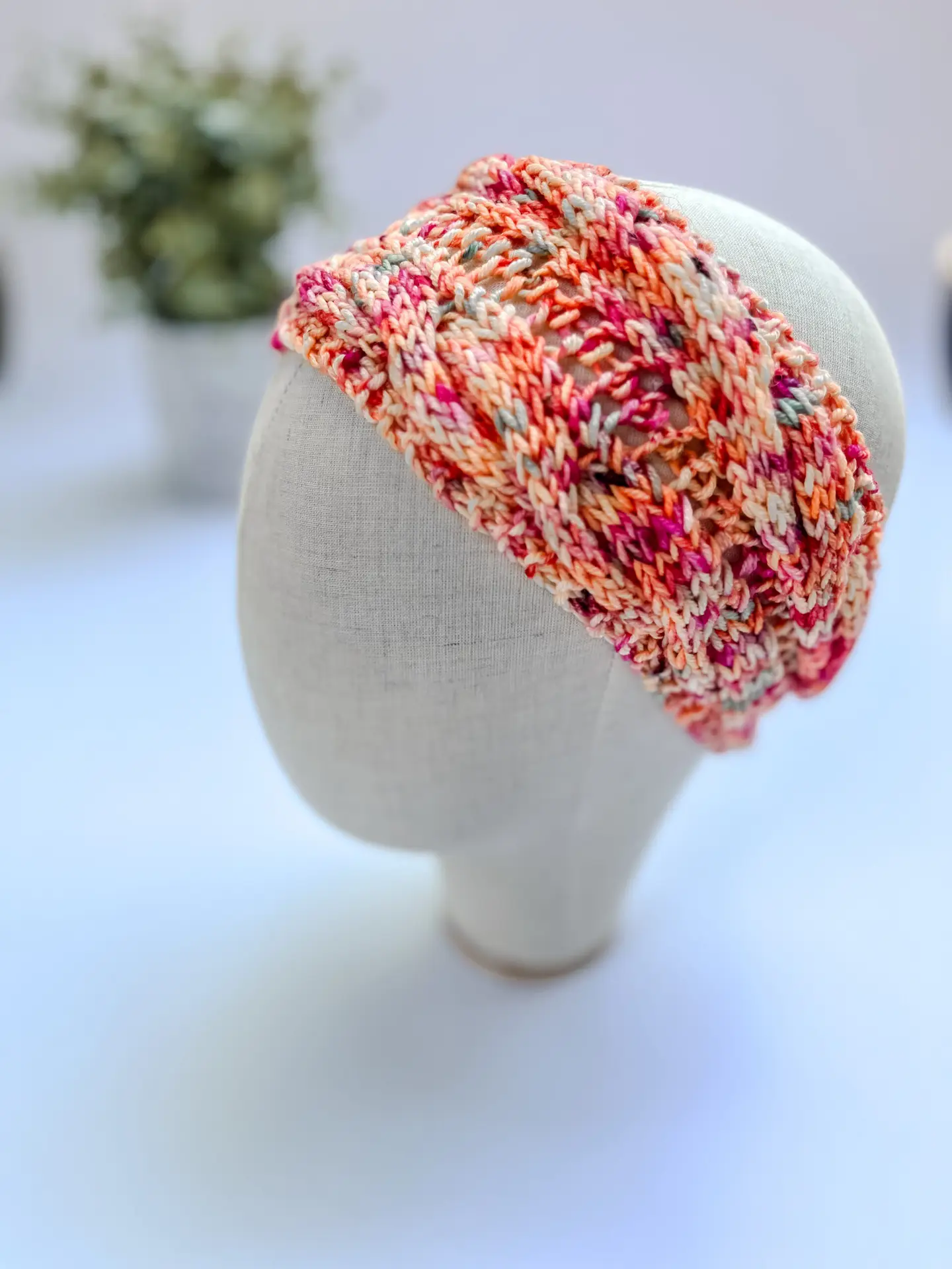 A bright hand-dyed merino ear warmer headband in a variety of oraanges, pinks, corals, and salmons is displayed on a cloth mannequin head.