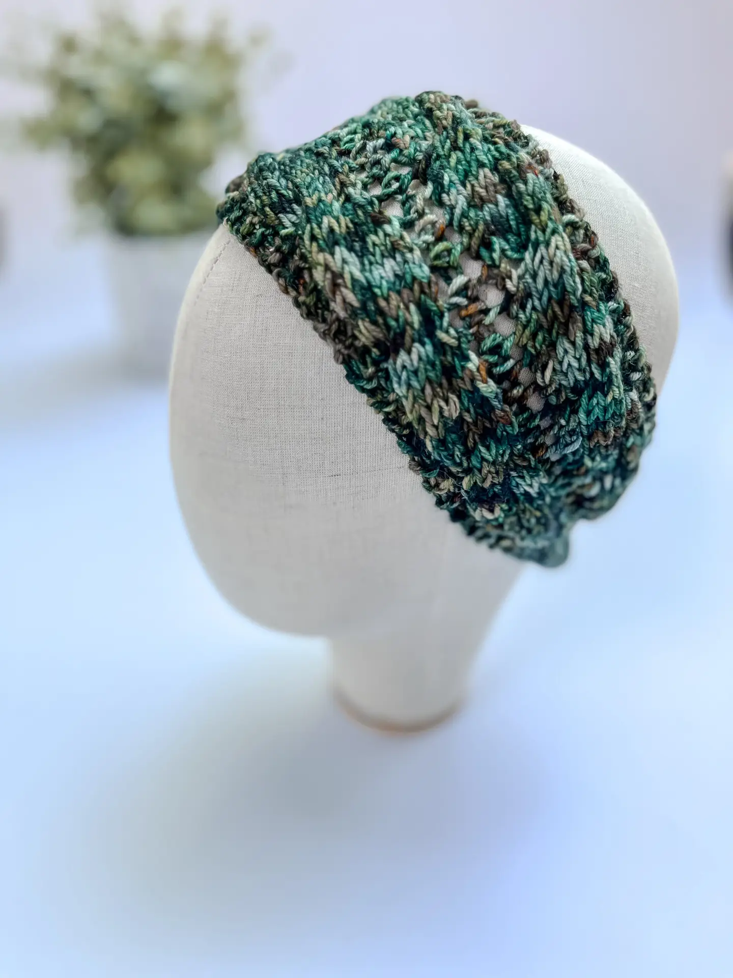 A green, blue and gold hand-dyed merino ear warmer headband is displayed on a cloth mannequin head.