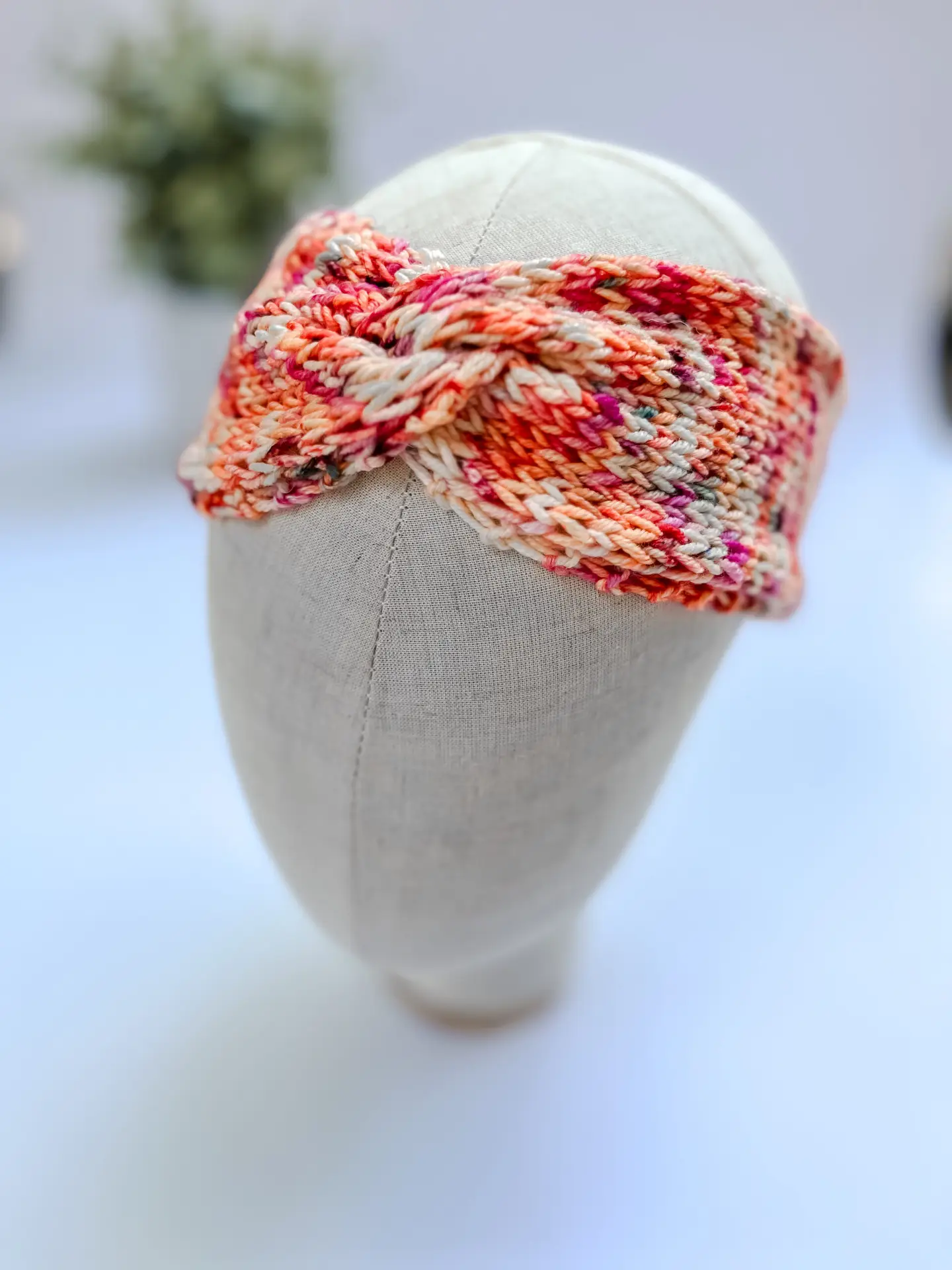 A coral, hand-dyed merino, twisted ear warmer headband is displayed on a cloth mannequin head.
