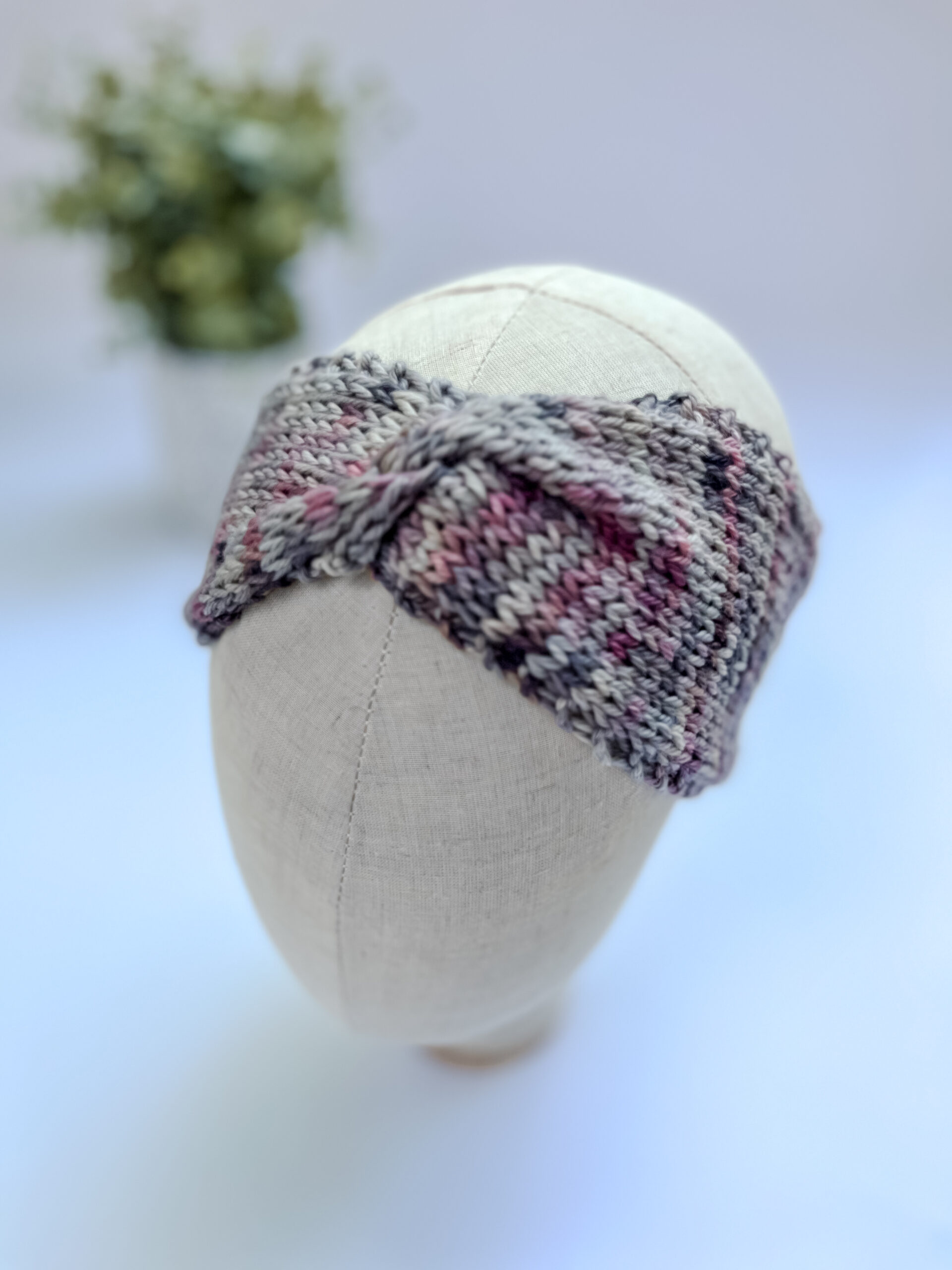 A gray, pink, & purple hand-dyed merino, twisted ear warmer headband is displayed on a cloth mannequin head.