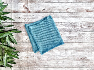 A pair of blue Virginia-grown cotton washcloths rests on a wood plank with greenery on the left border