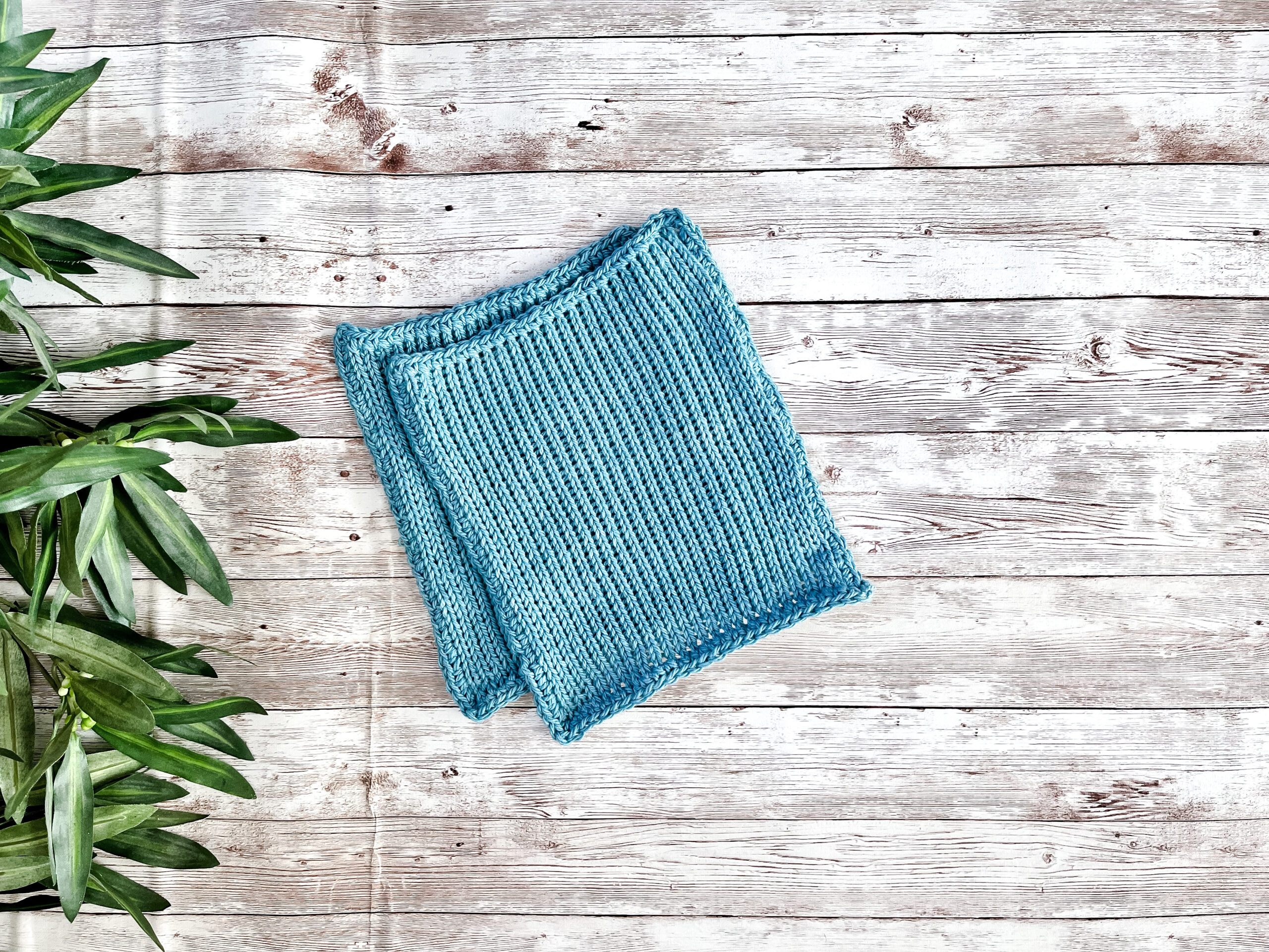 A pair of blue Virginia-grown cotton washcloths rests on a wood plank with greenery on the left border