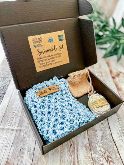 A black box holds a blue recycled cotton mesh produce tote, a tan bamboo/cotton soap saver, and a set of 5 Virginia-grown cotton reusable facial rounds. The box is on a wood panel with greenery in the background