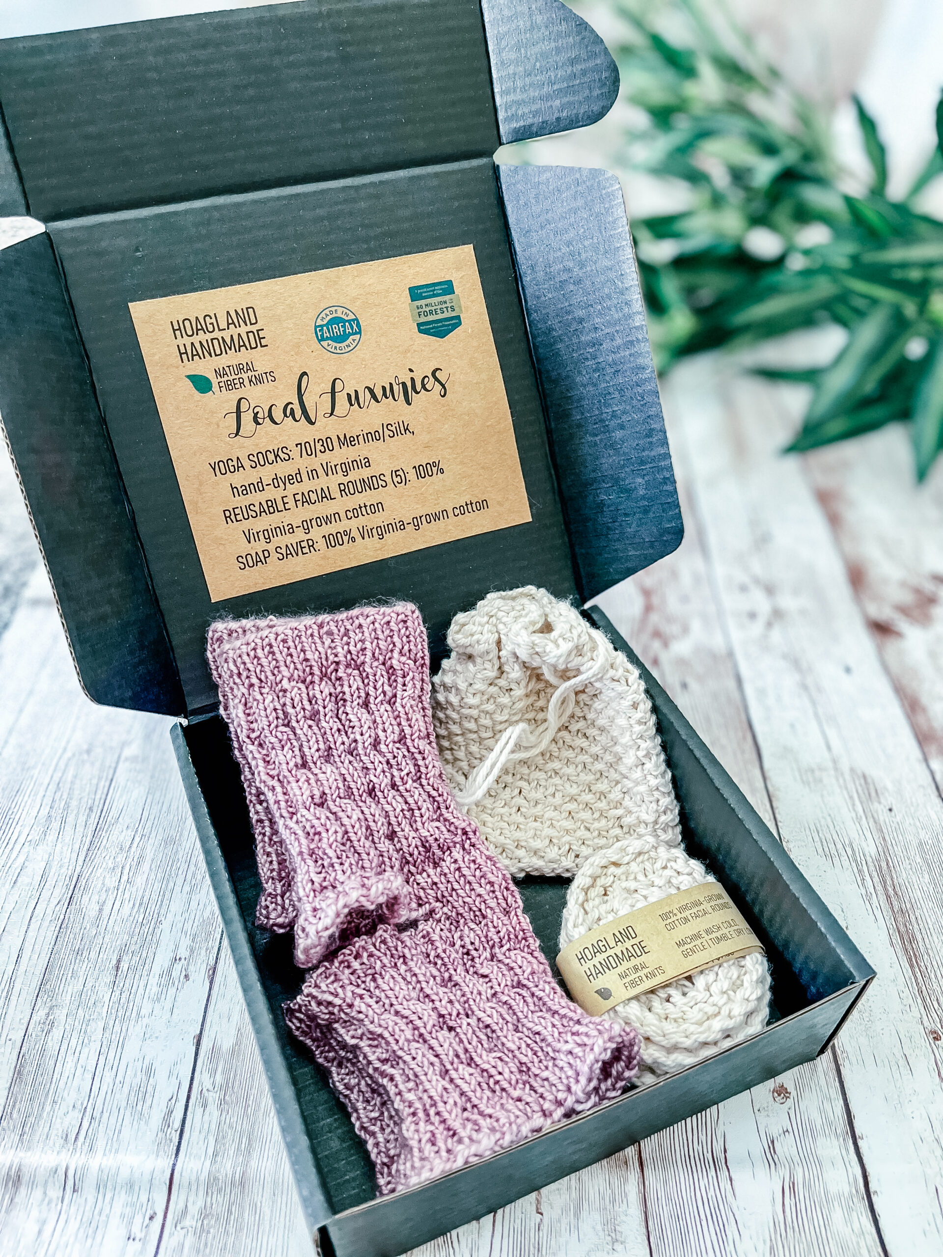 A black box contains a pair of hand-dyed merino/silk pink yoga socks, a Virginia-grown cotton soap saver and set of reusable facial rounds. The box sits on a wood panel with greenery in the background