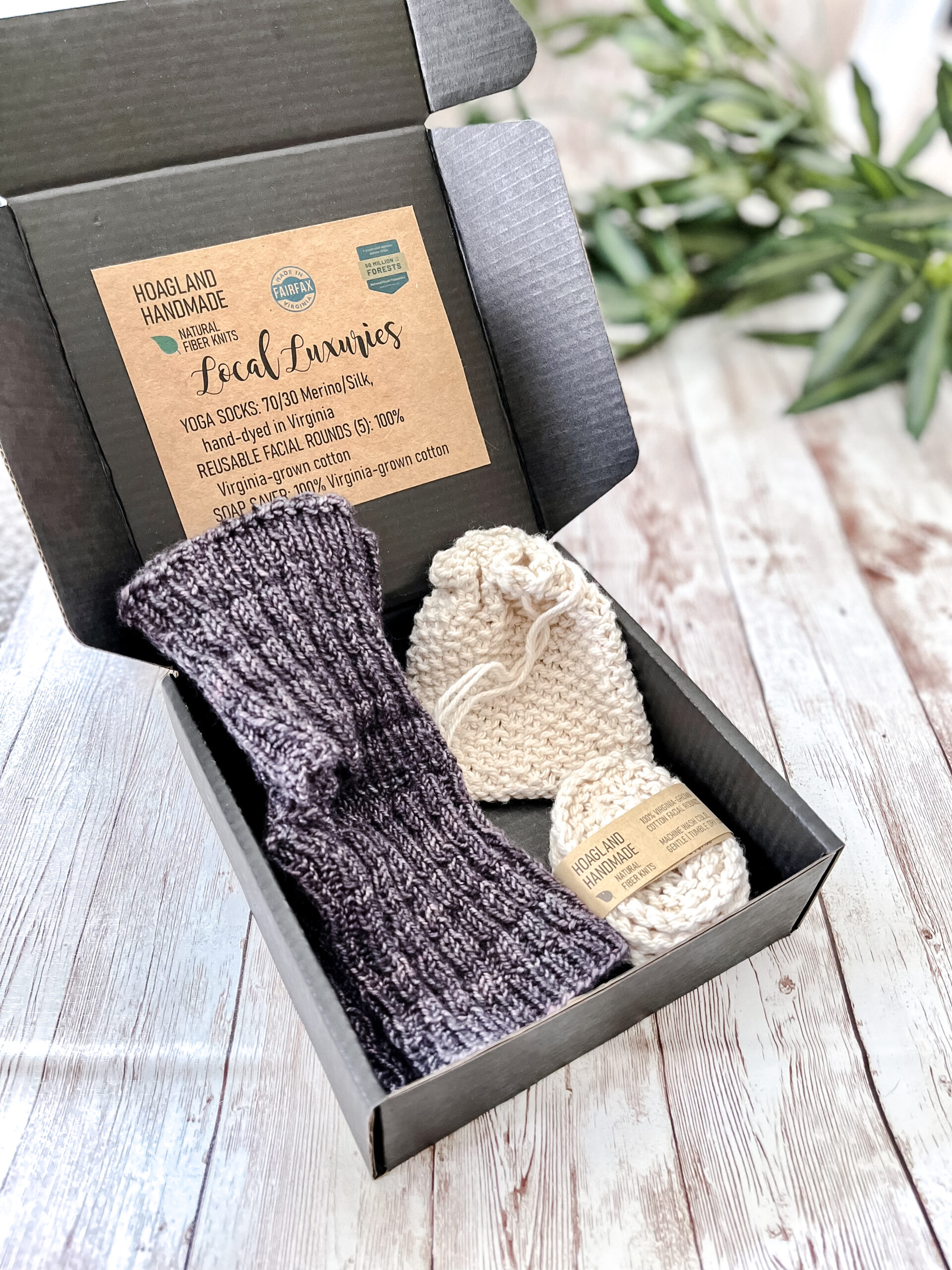 A black box contains a pair of hand-dyed merino/silk purplish gray & purple yoga socks, a Virginia-grown cotton soap saver and set of reusable facial rounds. The box sits on a wood panel with greenery in the background