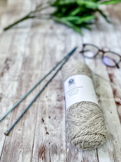 A natural light gray skein of yarn rests on a wood plank, with a pair of knitting needles, reading glasses, and greenery around it