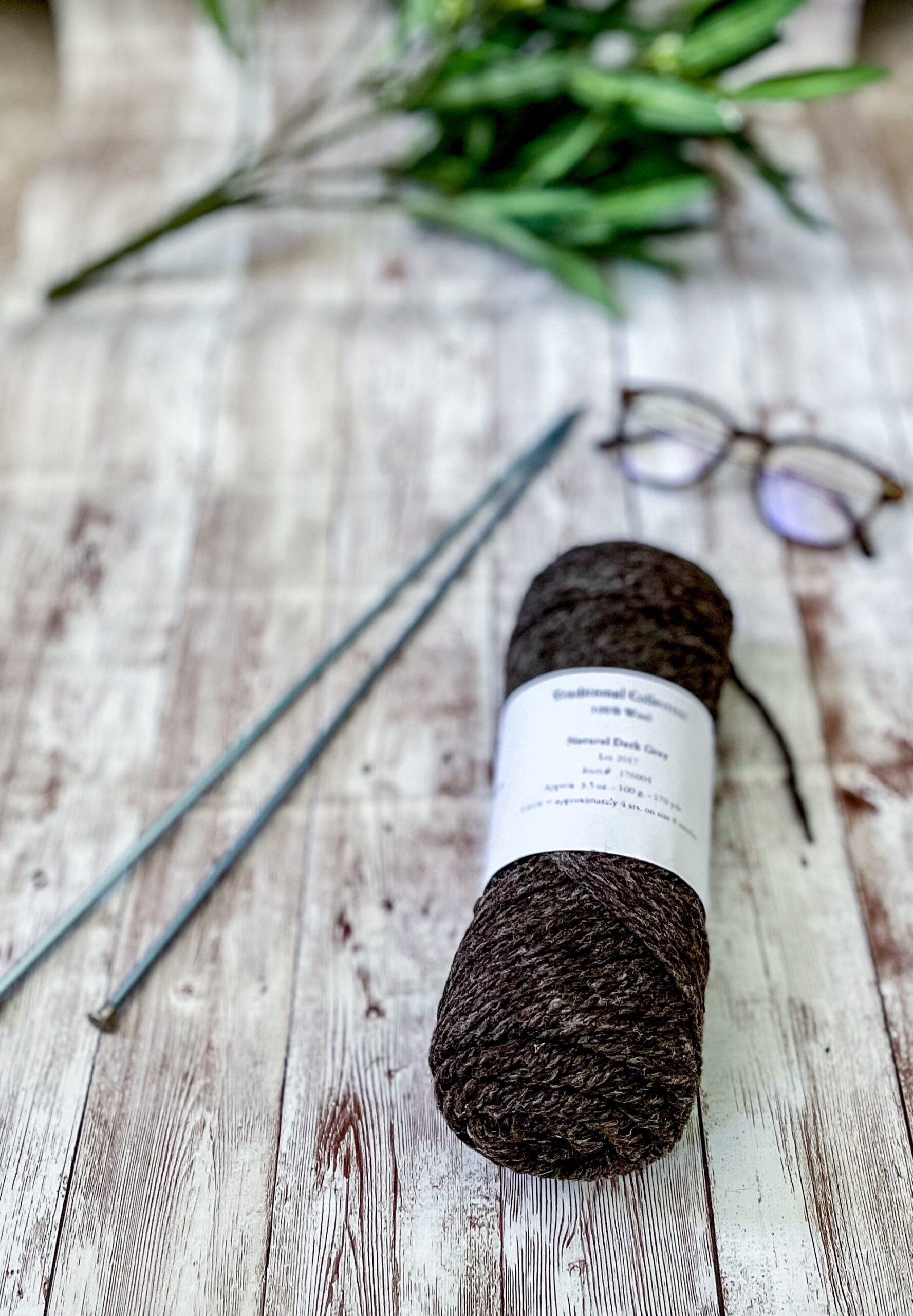 A natural dark gray skein of yarn rests on a wood plank, with a pair of knitting needles, reading glasses, and greenery around it