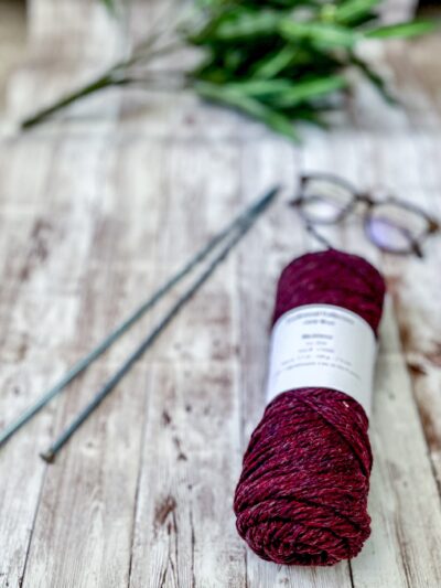 A deep purplish/red skein of yarn rests on a wood plank, with a pair of knitting needles, reading glasses, and greenery around it