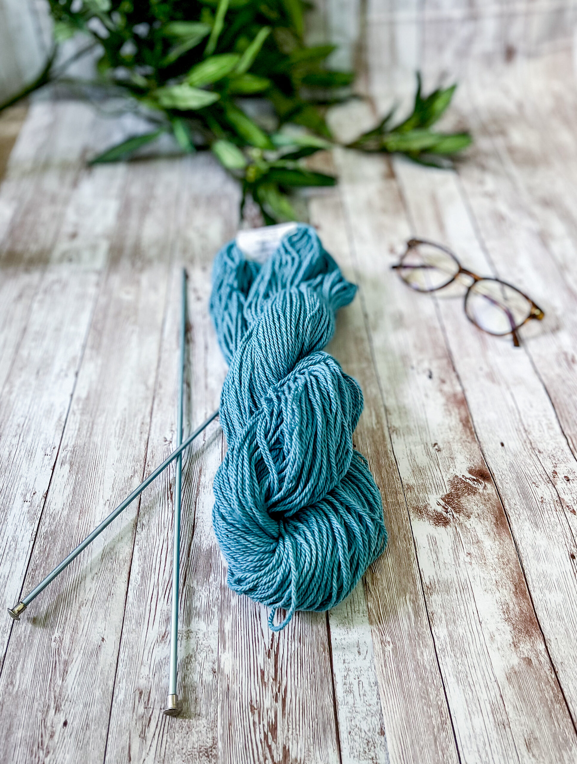 A skein of blue Virginia-grown cotton rests on a wood panel, with a pair of knitting needles, reading glasses and greenery around it