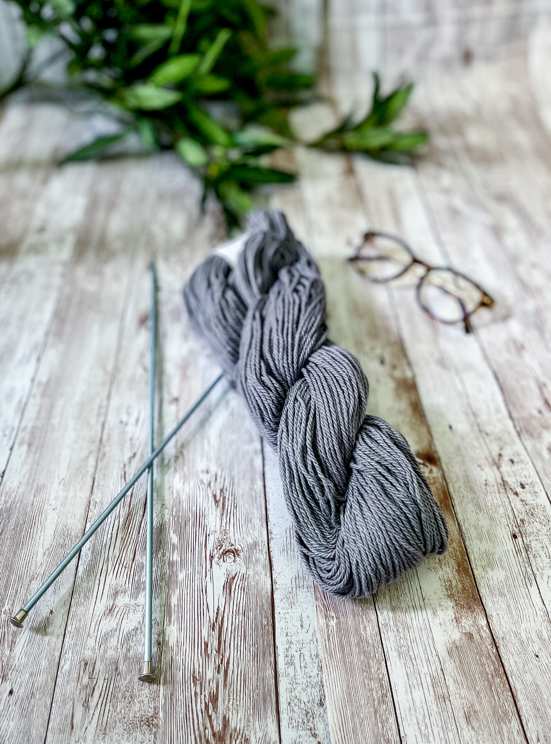 A skein of gray Virginia-grown cotton rests on a wood panel, with a pair of knitting needles, reading glasses and greenery around it