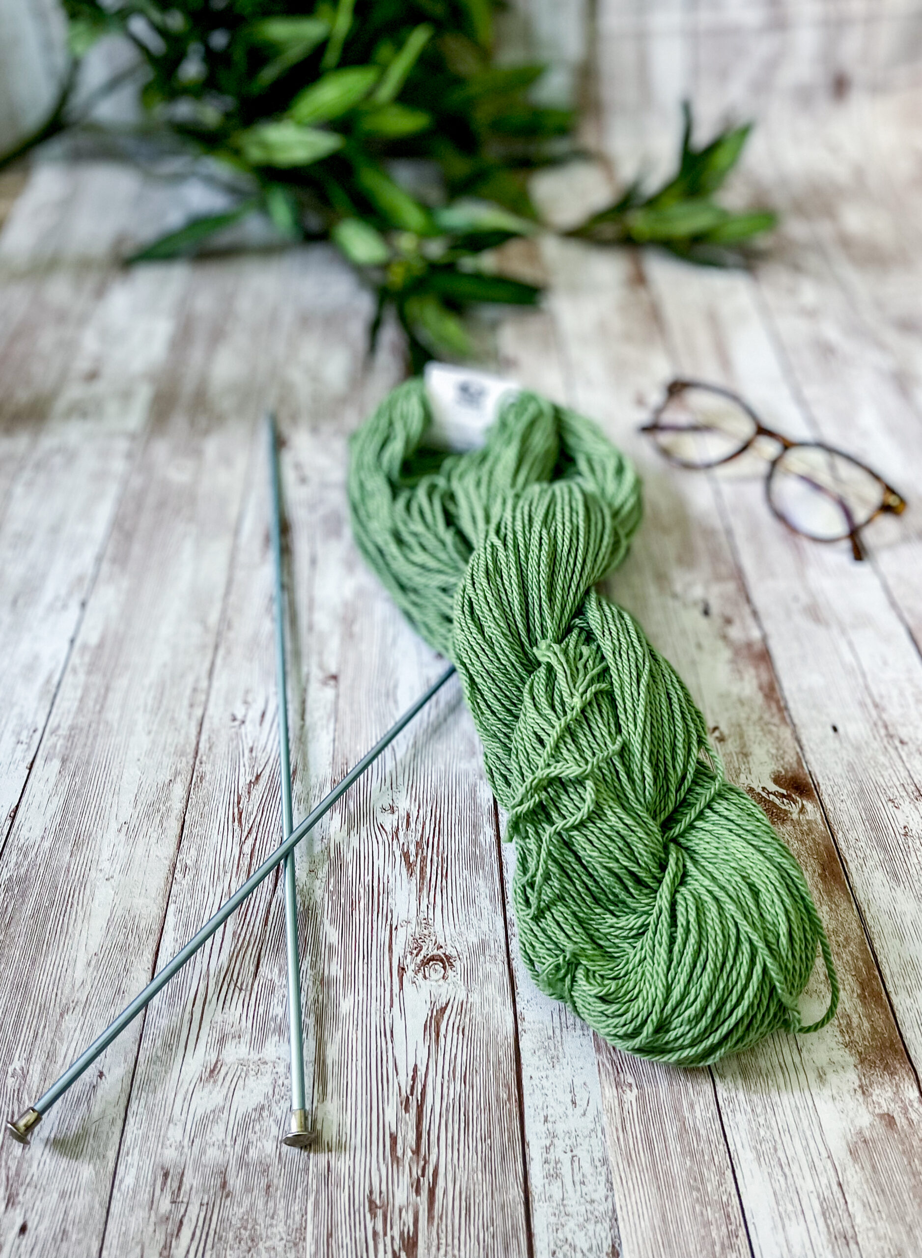 A skein of green Virginia-grown cotton rests on a wood panel, with a pair of knitting needles, reading glasses and greenery around it