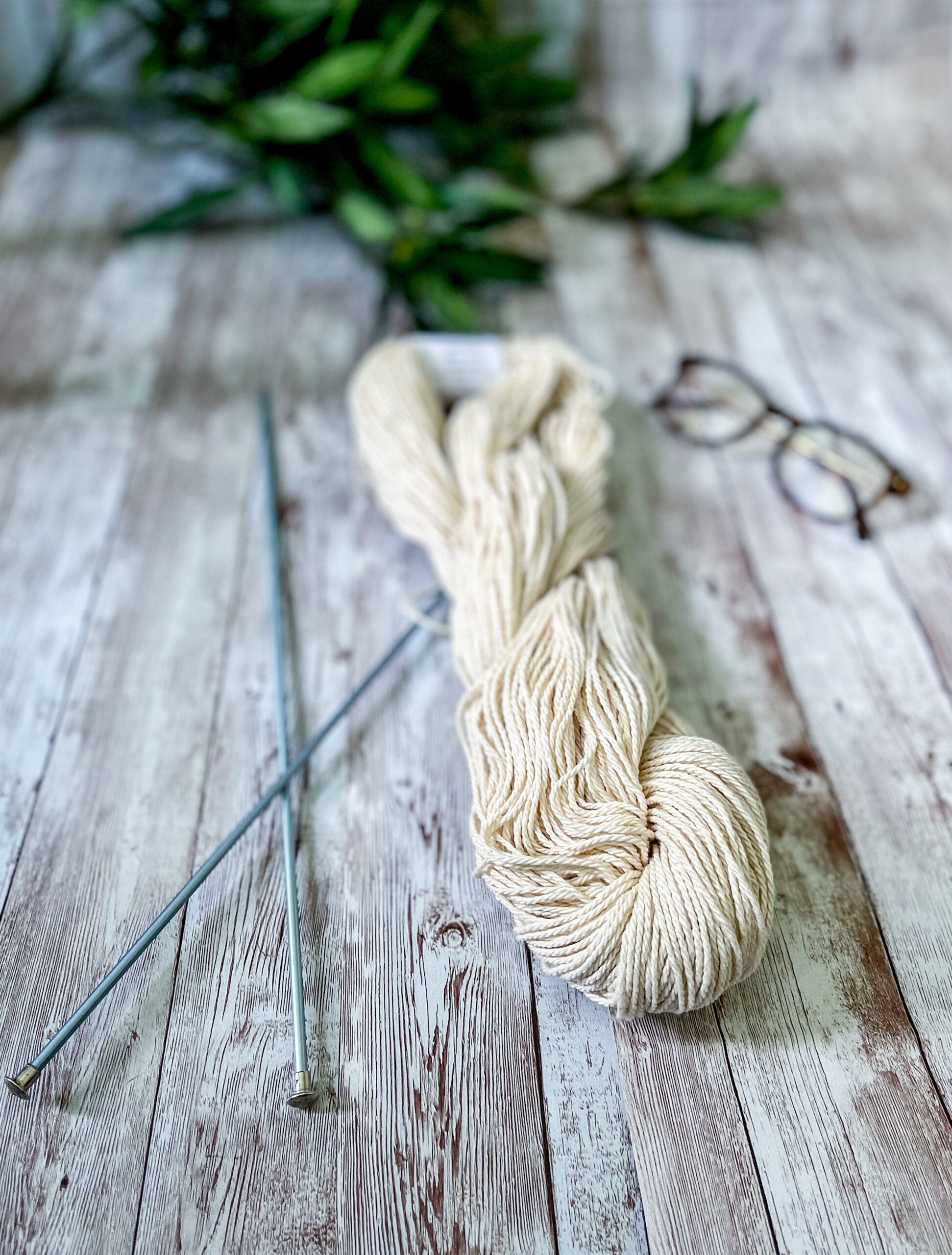 A skein of undyed, off-white Virginia-grown cotton rests on a wood panel, with a pair of knitting needles, reading glasses and greenery around it