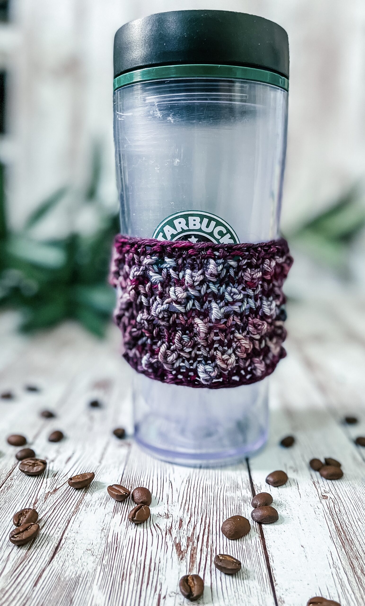 A tall coffee tumbler sports a purple and teal hand-dyed merino coffee sleeve with diagonal bobbles. It sits on a wood plank with coffee beans scattered around and greenery in the background