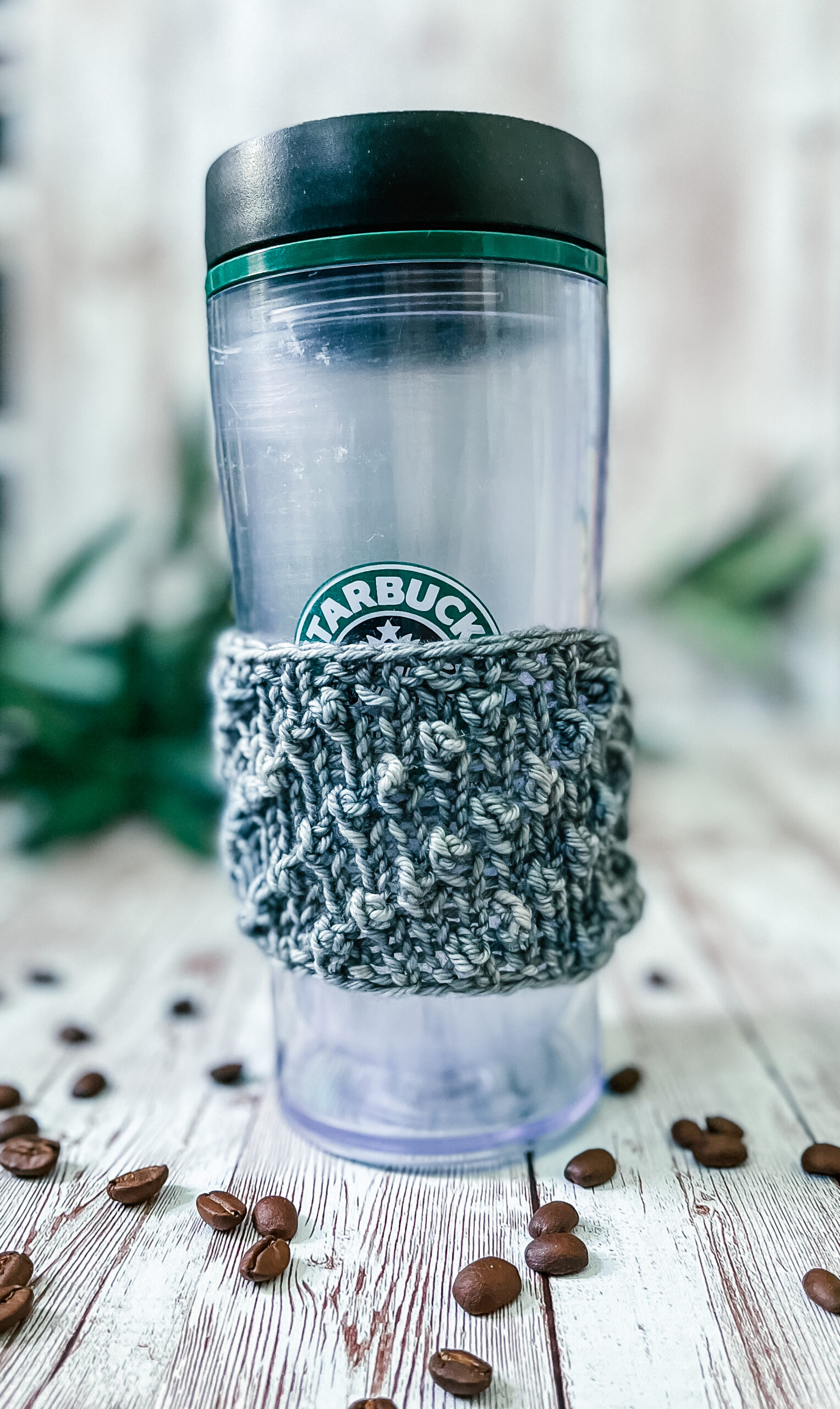 A tall coffee tumbler sports a gray hand-dyed merino coffee sleeve with diagonal bobbles. It sits on a wood plank with coffee beans scattered around and greenery in the background