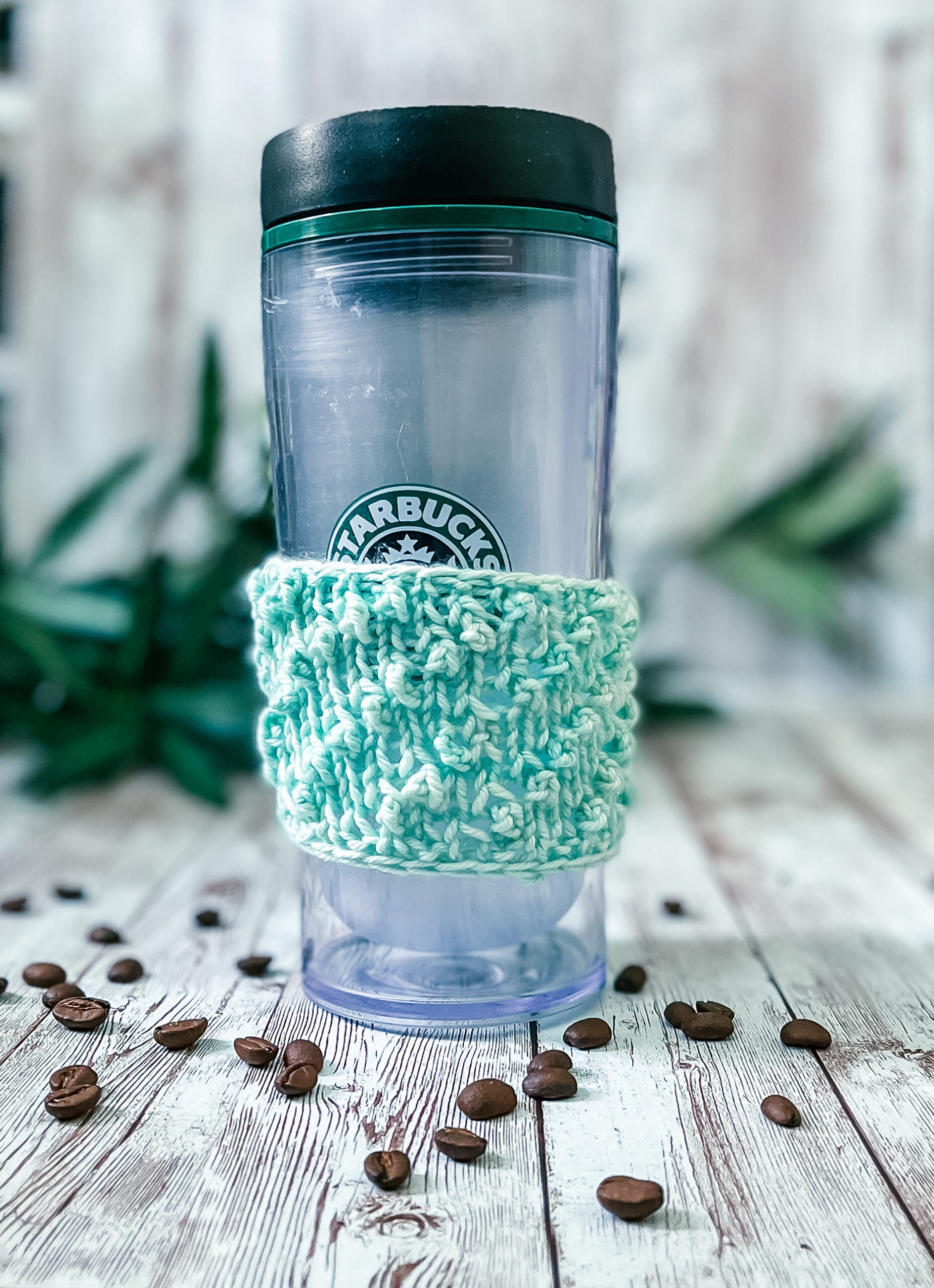 A tall coffee tumbler sports a pale greenhand-dyed merino coffee sleeve with diagonal bobbles. It sits on a wood plank with coffee beans scattered around and greenery in the background