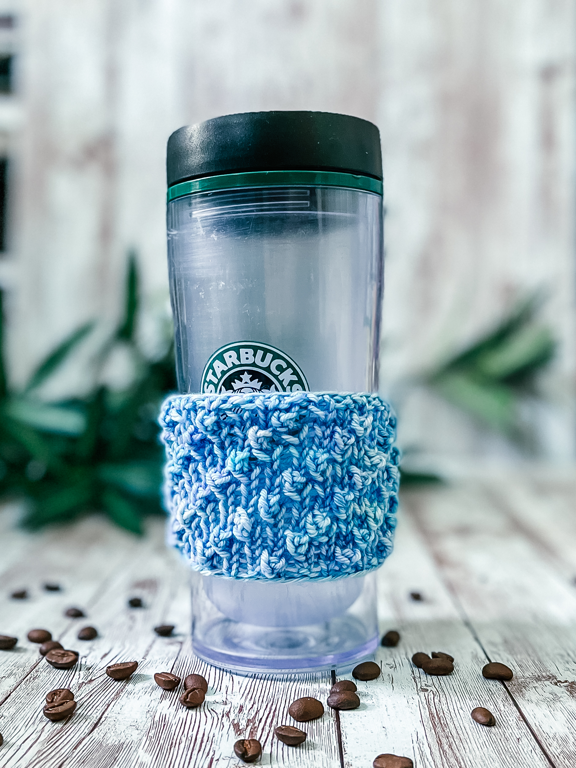 A tall coffee tumbler sports a blue hand-dyed merino coffee sleeve with diagonal bobbles. It sits on a wood plank with coffee beans scattered around and greenery in the background