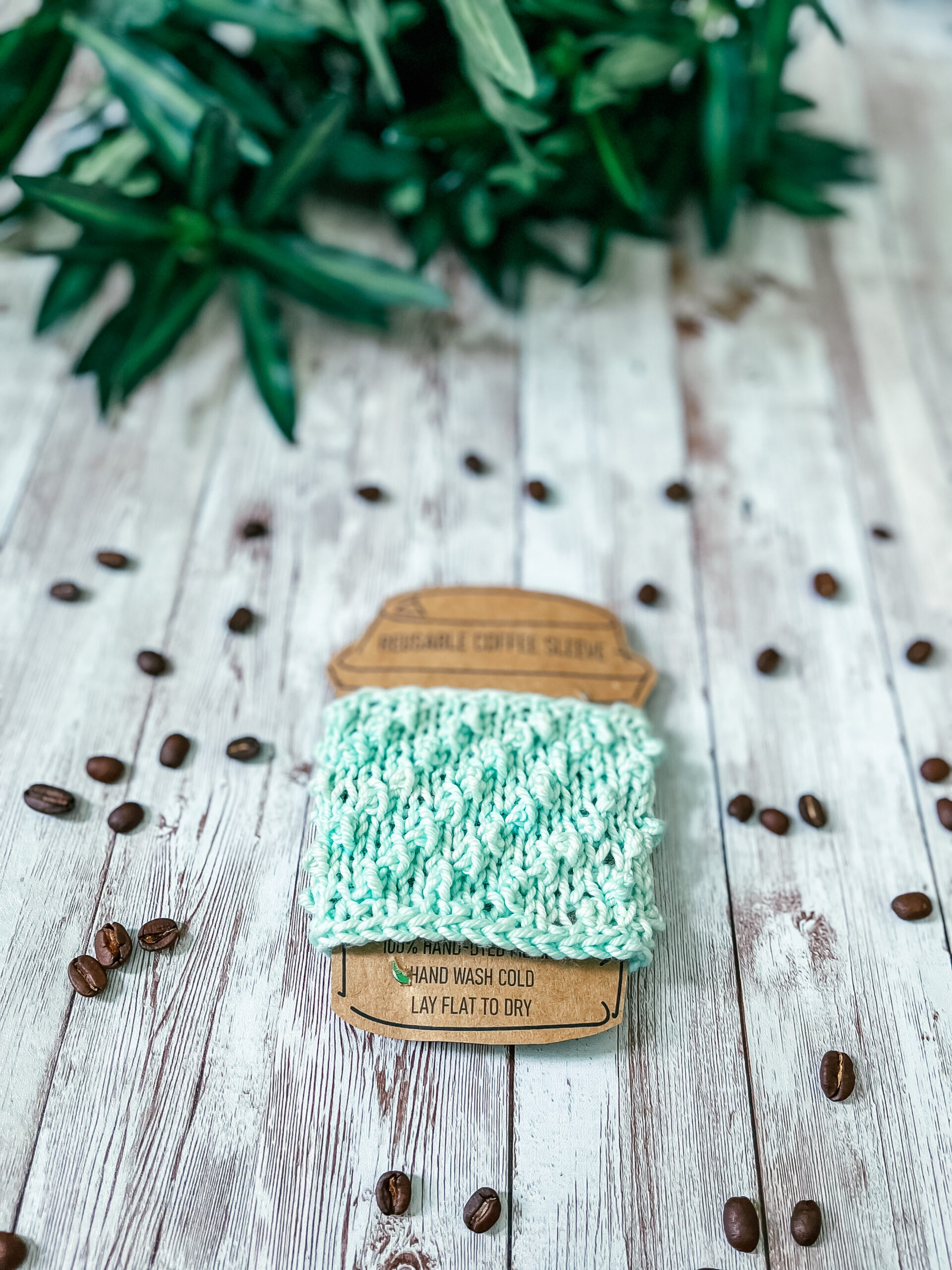 A light green coffee sleeve is wrapped around a kraft paper image of a coffee cup. It rests on a wood plank, surrounded by scattered coffee beans and greenery in the background