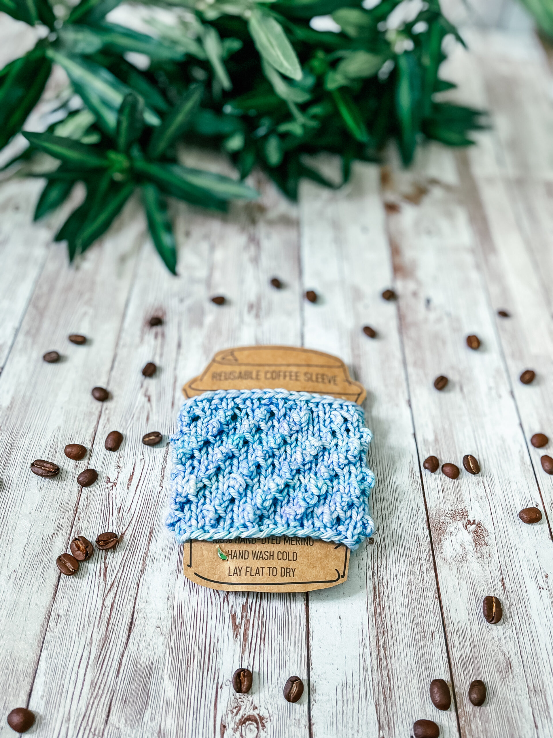 A light blue coffee sleeve is wrapped around a kraft paper image of a coffee cup. It rests on a wood plank, surrounded by scattered coffee beans and greenery in the background