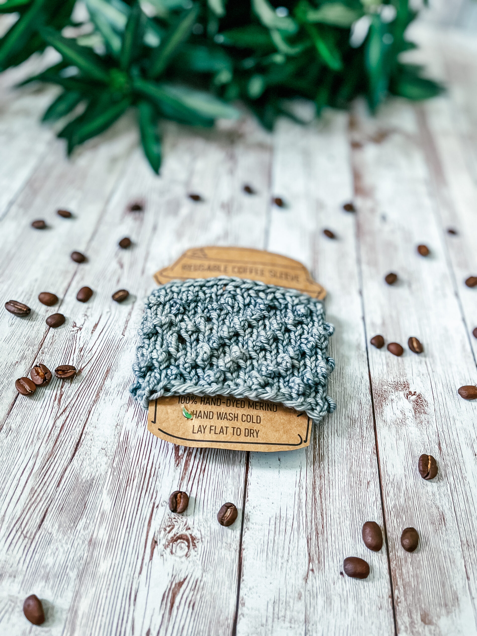 A light gray coffee sleeve is wrapped around a kraft paper image of a coffee cup. It rests on a wood plank, surrounded by scattered coffee beans and greenery in the background