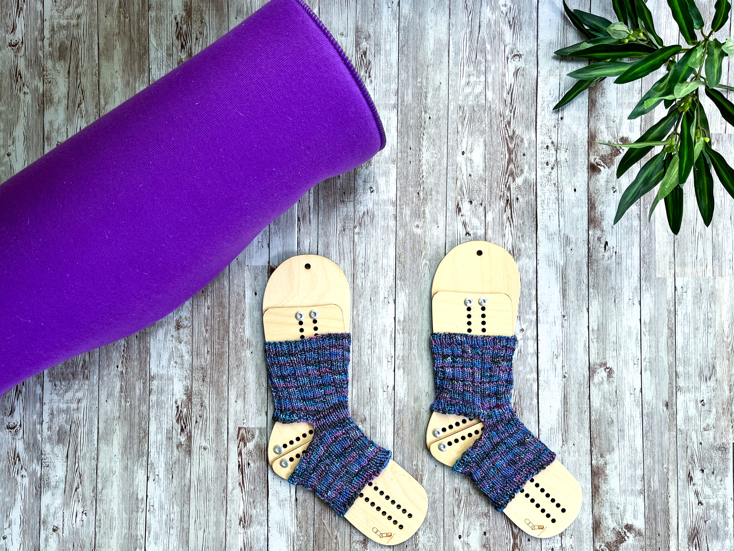 A pair of purple and blue yoga socks on wooden sock blockers are surrounded by a rolled up yoga mat and greenery on a wood panel
