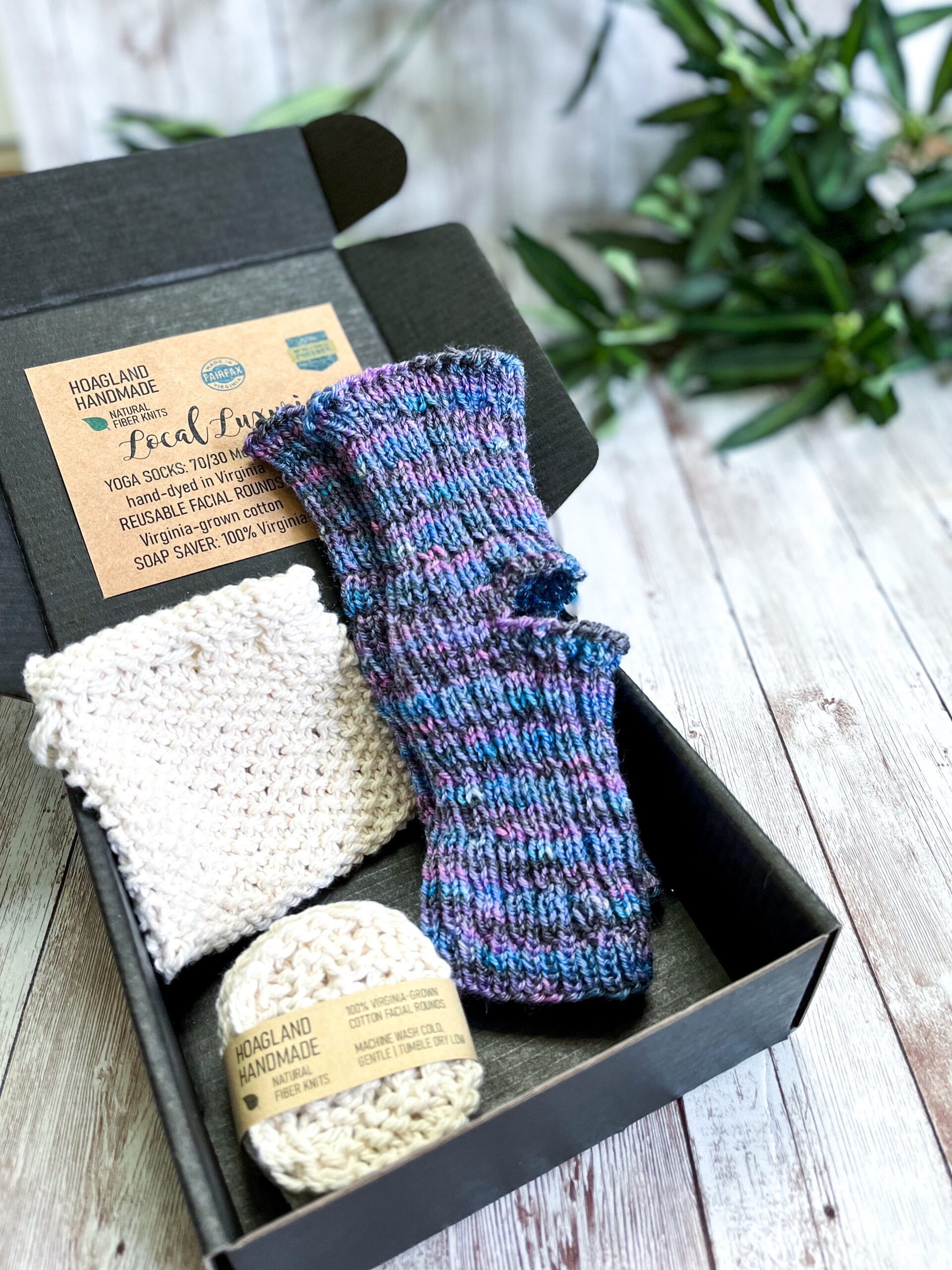 A black box contains a pair of hand-dyed merino blue & purple yoga socks, a Virginia-grown cotton soap saver and set of reusable facial rounds. The box sits on a wood panel with greenery in the background