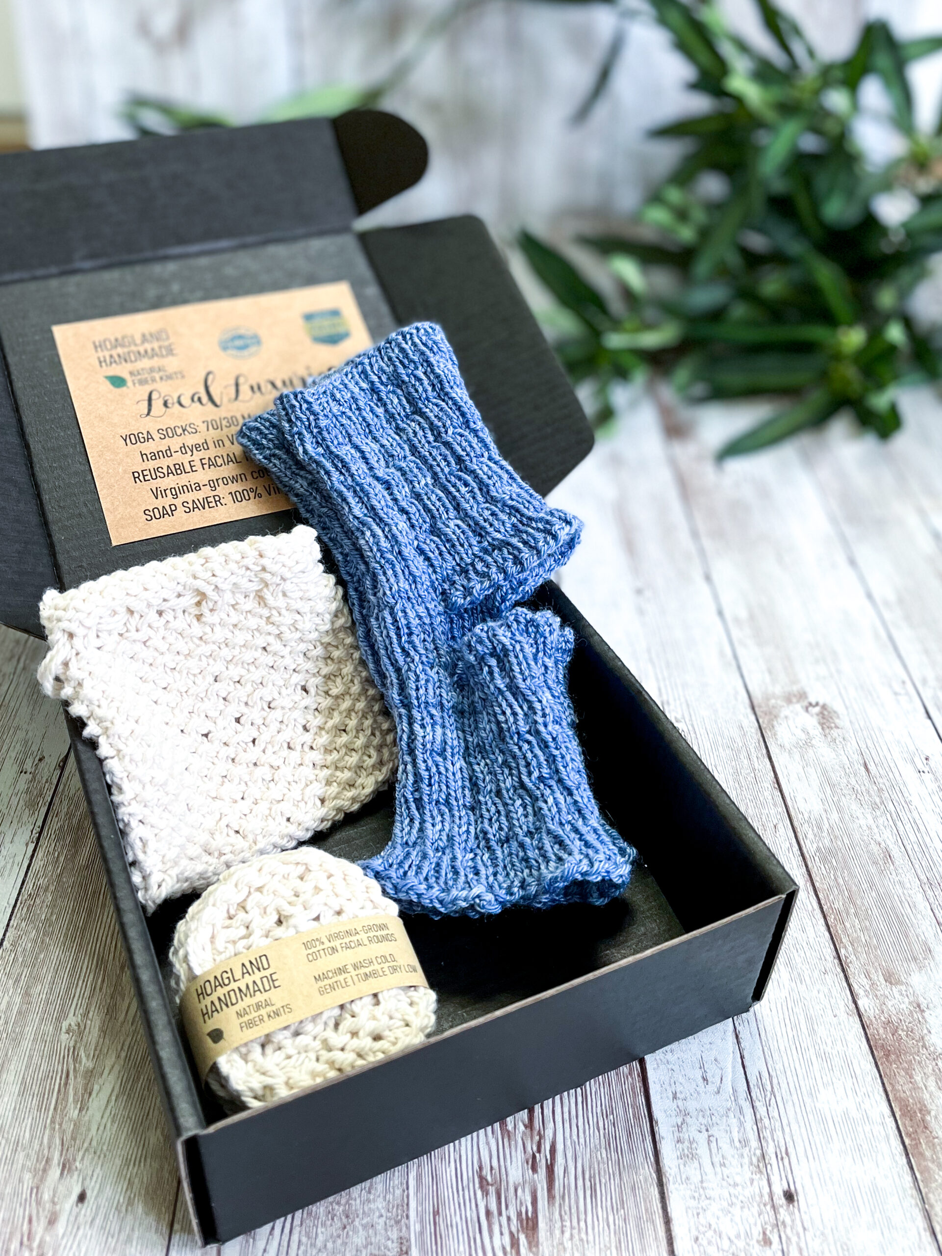 A black box contains a pair of hand-dyed merino/bamboo blue yoga socks, a Virginia-grown cotton soap saver and set of reusable facial rounds. The box sits on a wood panel with greenery in the background
