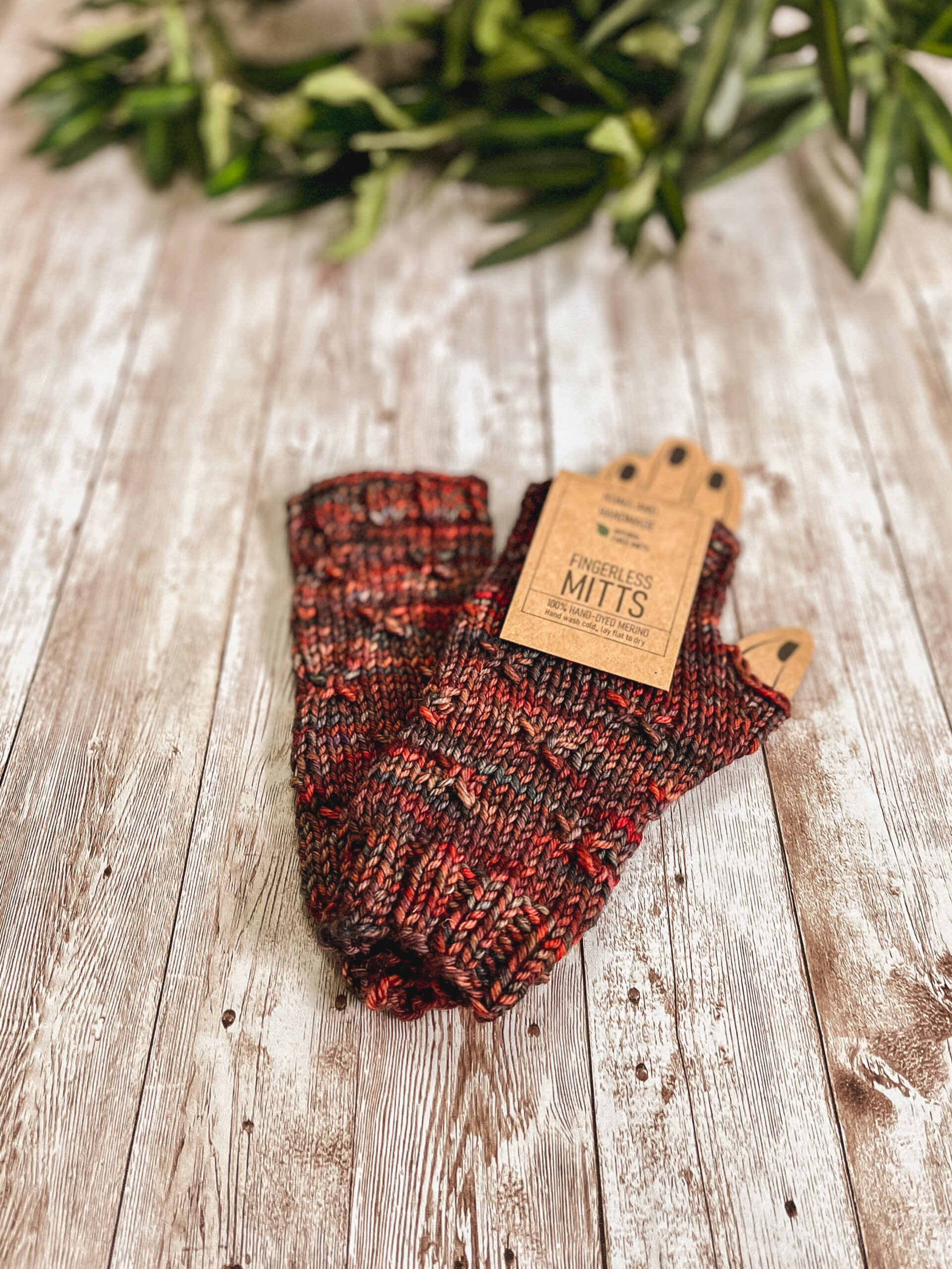 A pair of fingerless mitts in an autumn mix of colors lays on a wood panel with greenery in the background