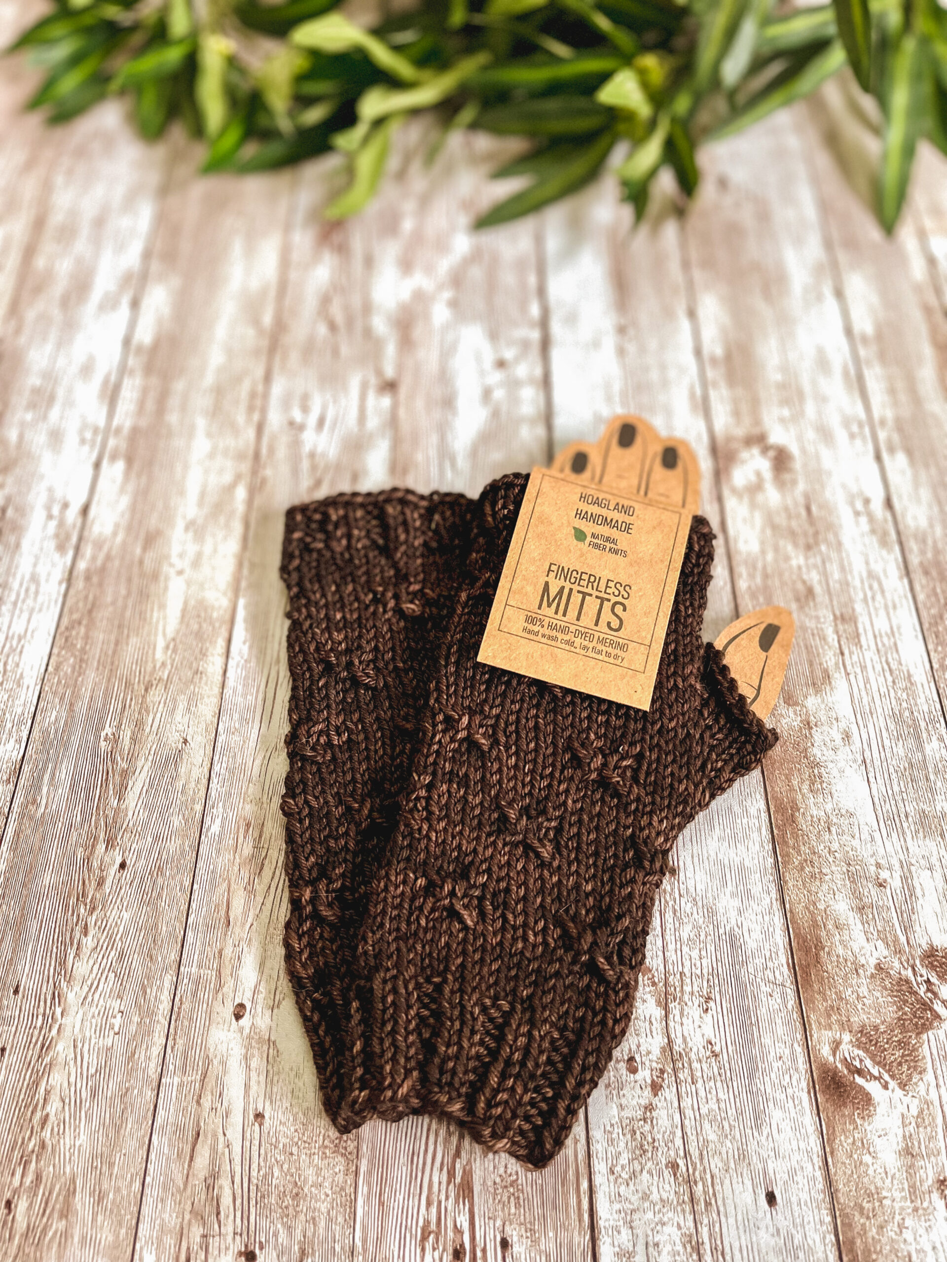 A pair of brown fingerless mitts with a decorative gathered pattern sit on a wood plank with greenery in the background. There is a tag at the top that reads Hoagland Handmade, Natural Fiber Knits, Fingerless Mitts 100% hand dyed merino