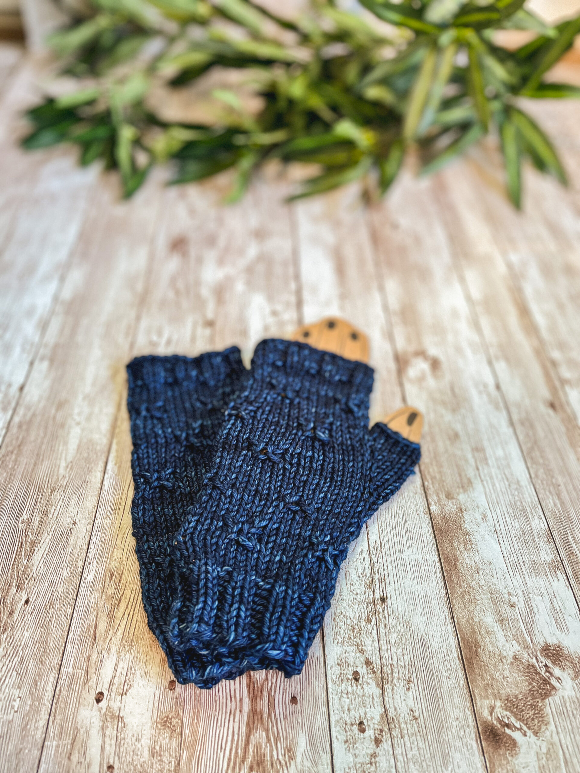A pair of blue fingerless mitts with a decorative gathered pattern sit on a wood plank with greenery in the background