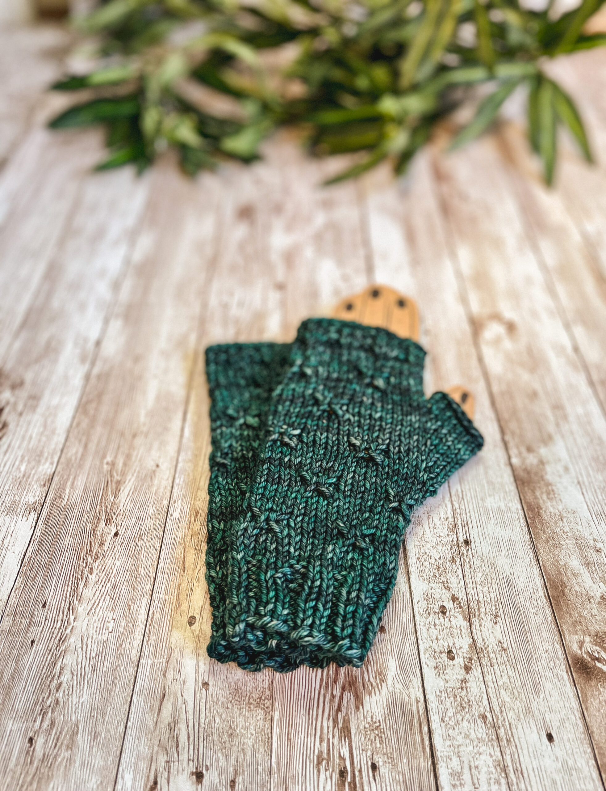 A pair of green fingerless mitts with a decorative gathered pattern sit on a wood plank with greenery in the background