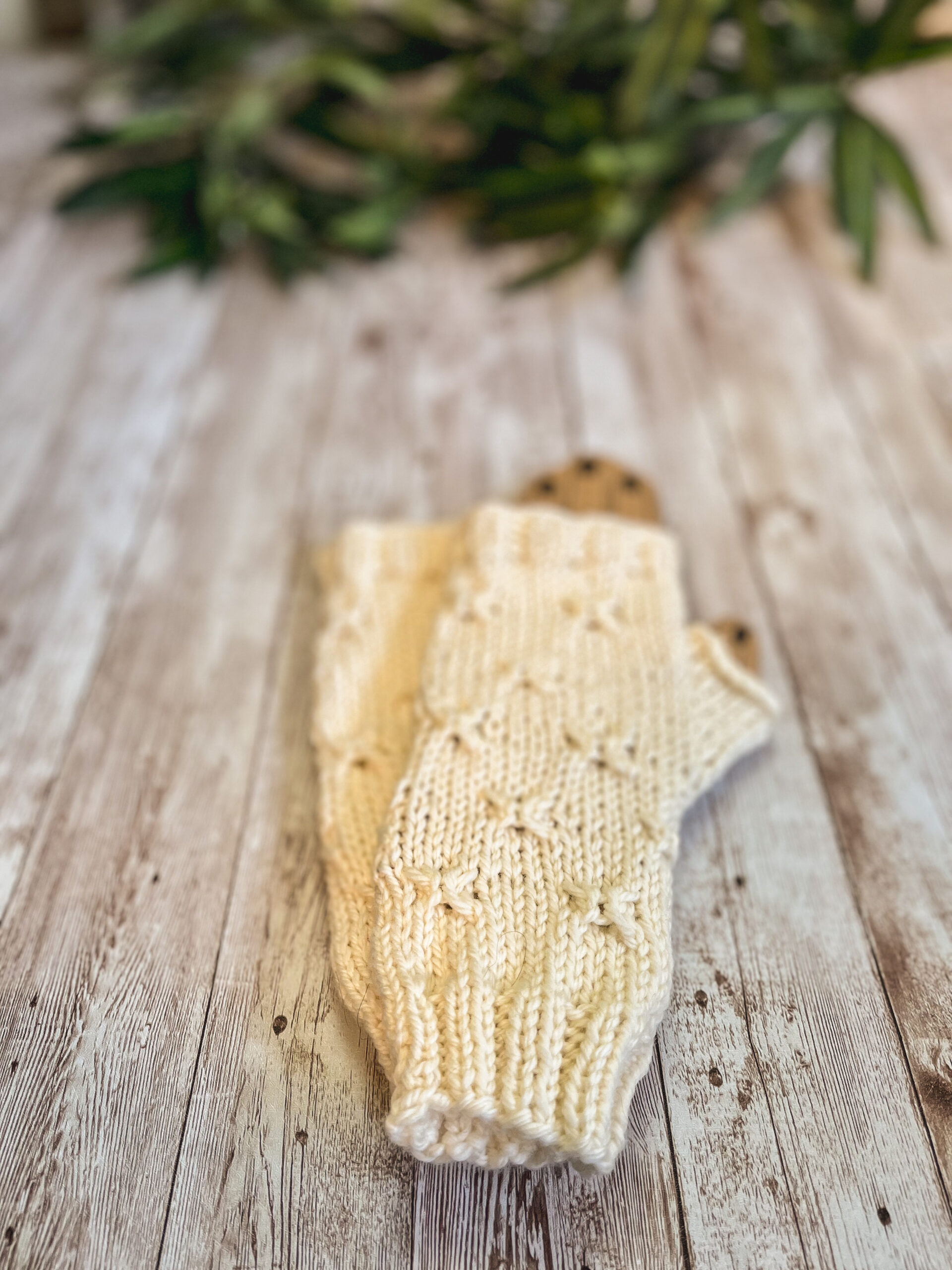 A pair of off white fingerless mitts with a decorative gathered pattern sit on a wood plank with greenery in the background