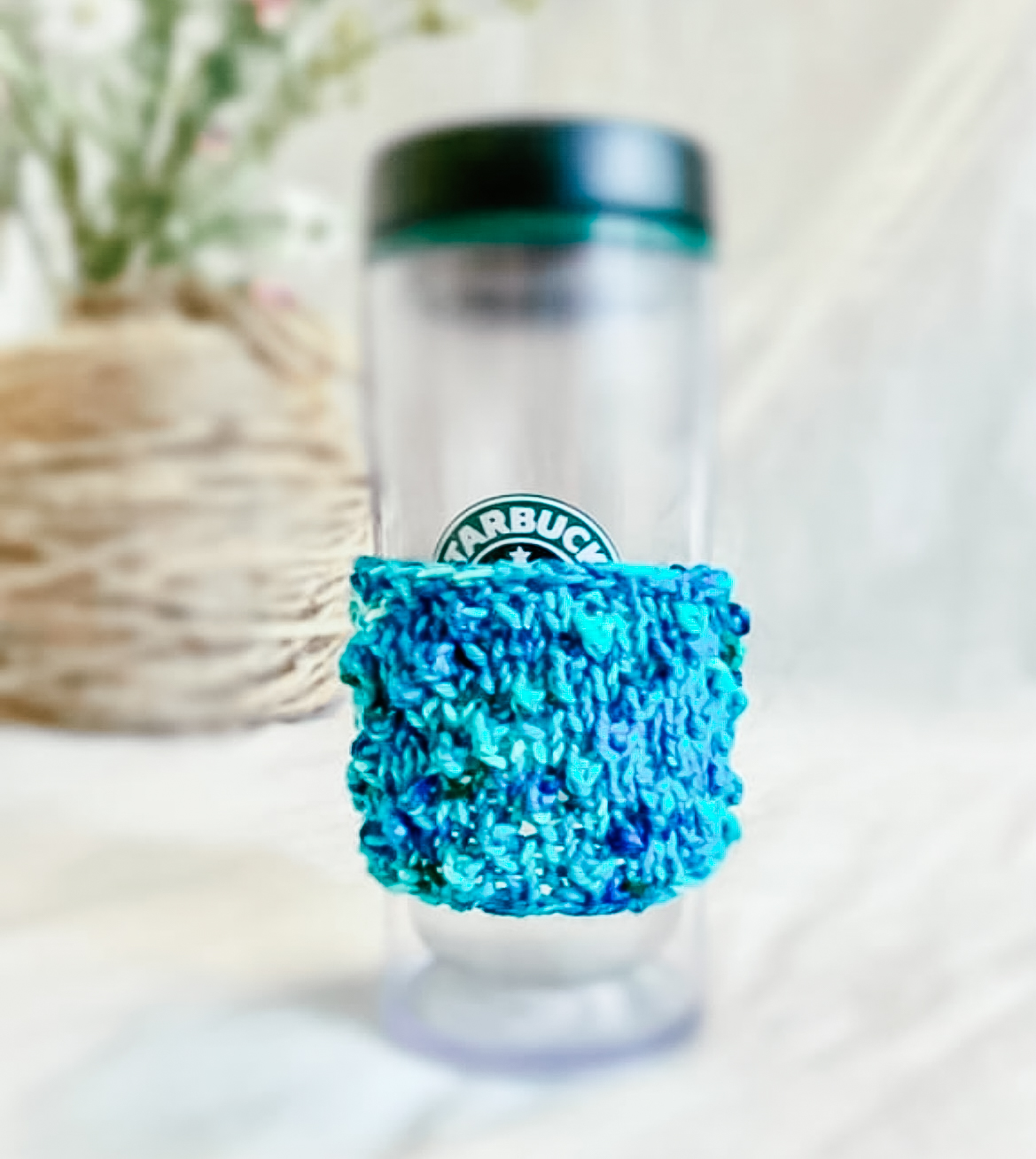 A tall coffee tumbler sports a teal hand-dyed merino coffee sleeve with diagonal bobbles. There is a floral arrangement in the background