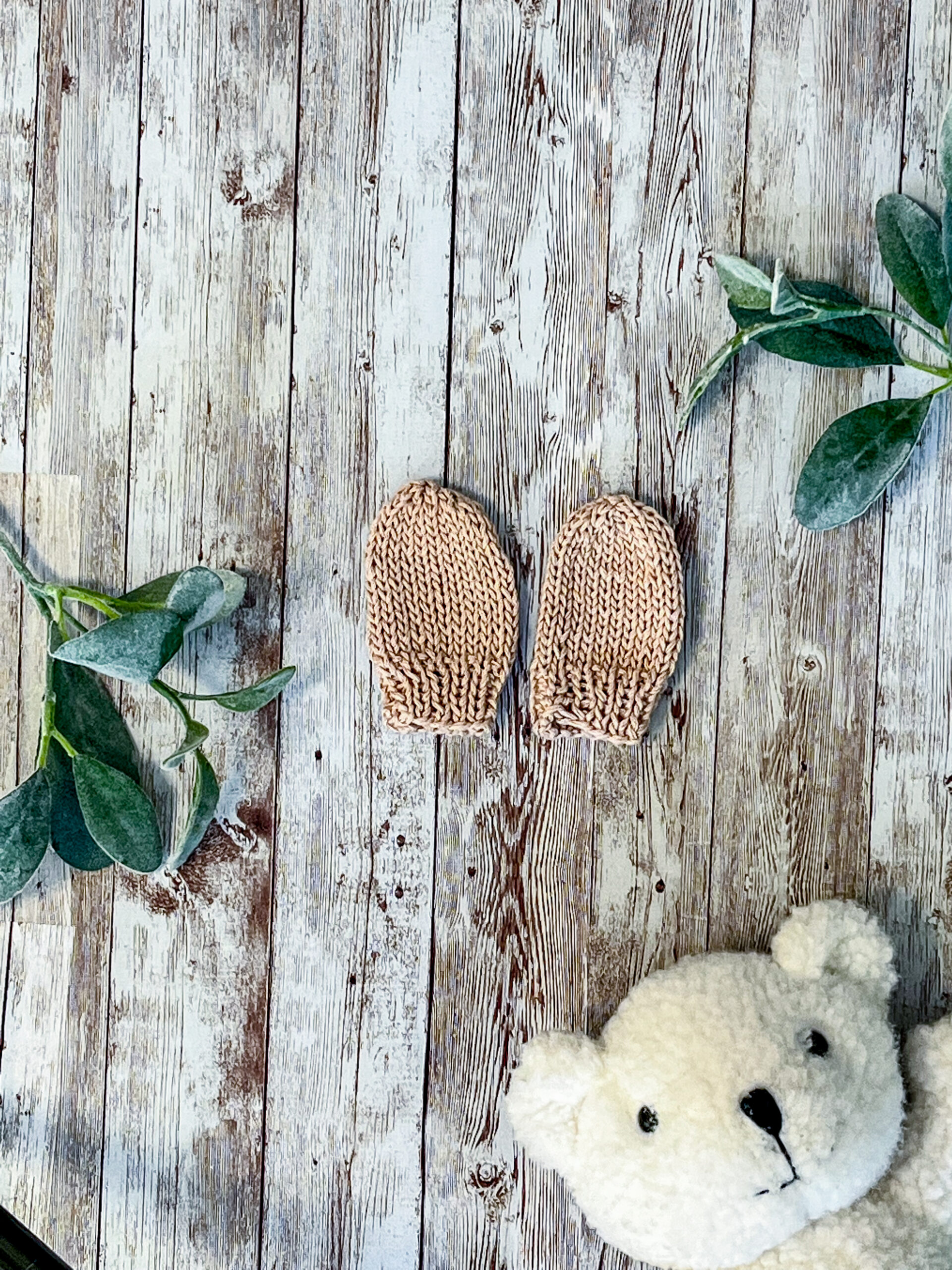 A pair of tan organic cotton hand-knit newborn mitts are on a wood background, with sage leaves and a white teddy bear face