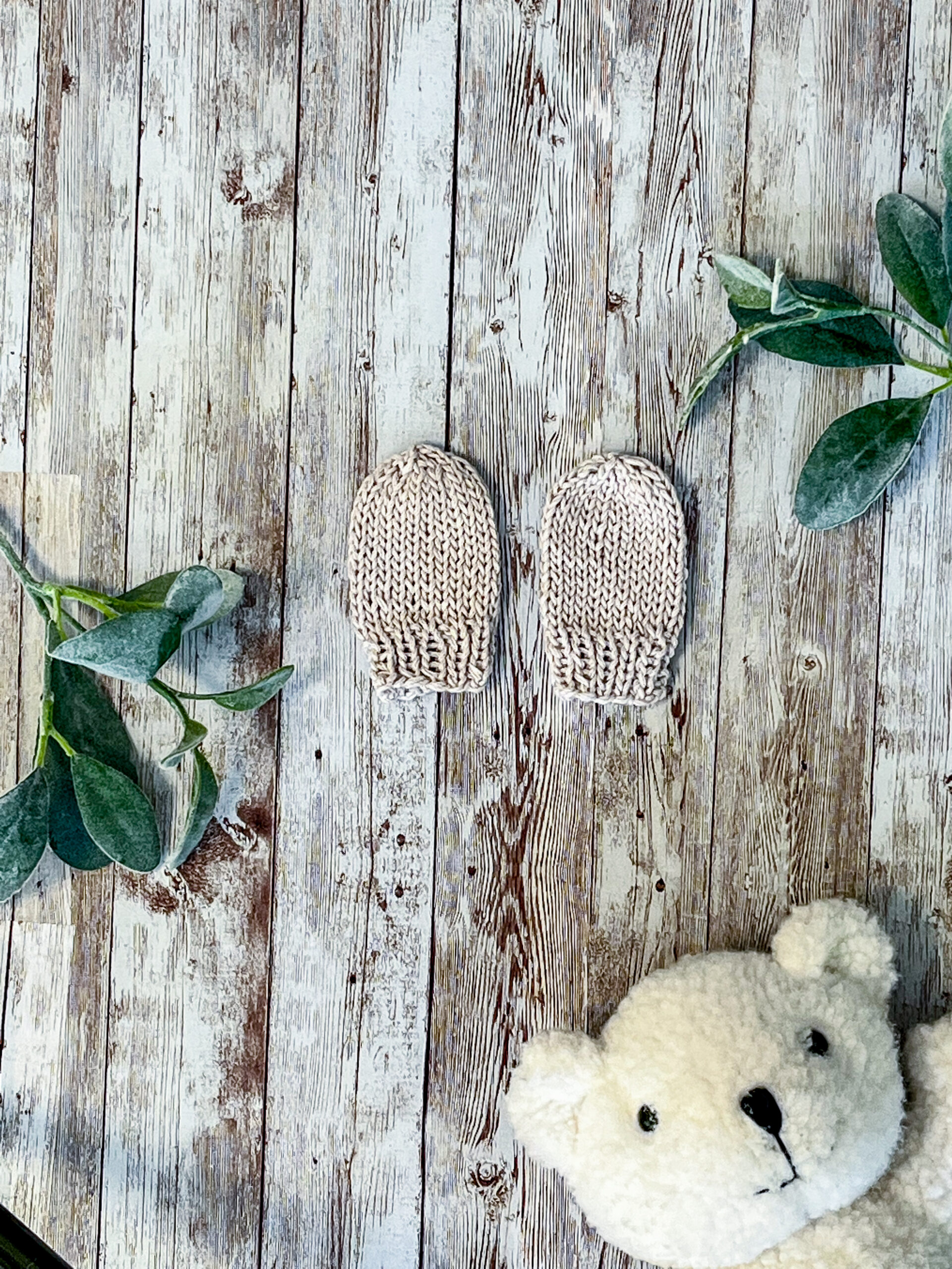 A pair of gray organic cotton hand-knit newborn mitts are on a wood background, with sage leaves and a white teddy bear face